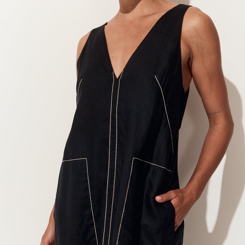 A close-up of a model wearing a black sleeveless V-neck linen jumpsuit with contrasting light beige stitching and large front patch pockets, set against a plain off-white wall.