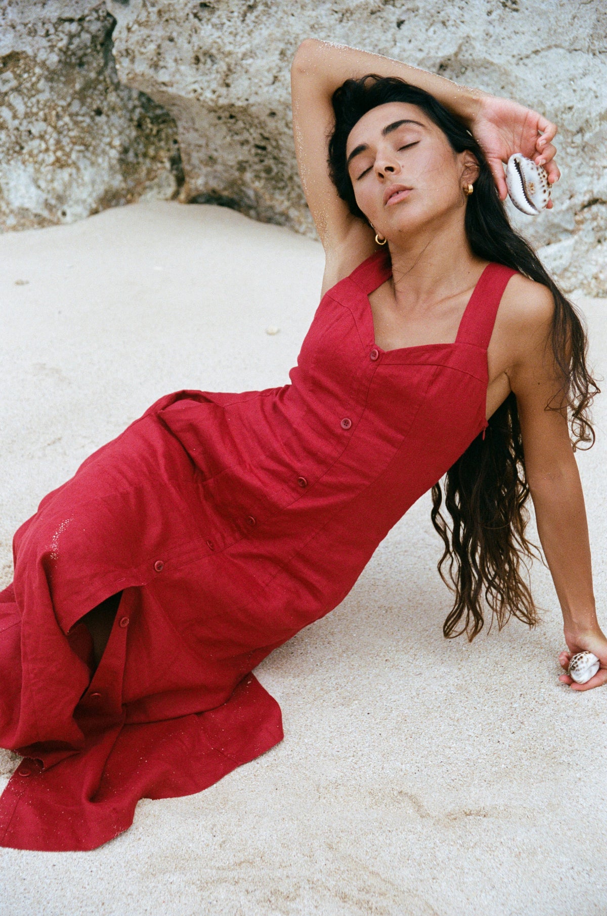 Eden Linen Midi Dress in Cherry Red - woman reclined on sand, sweetheart neckline and button-front placket, beach rocks backdrop - Lilly Pilly Collection