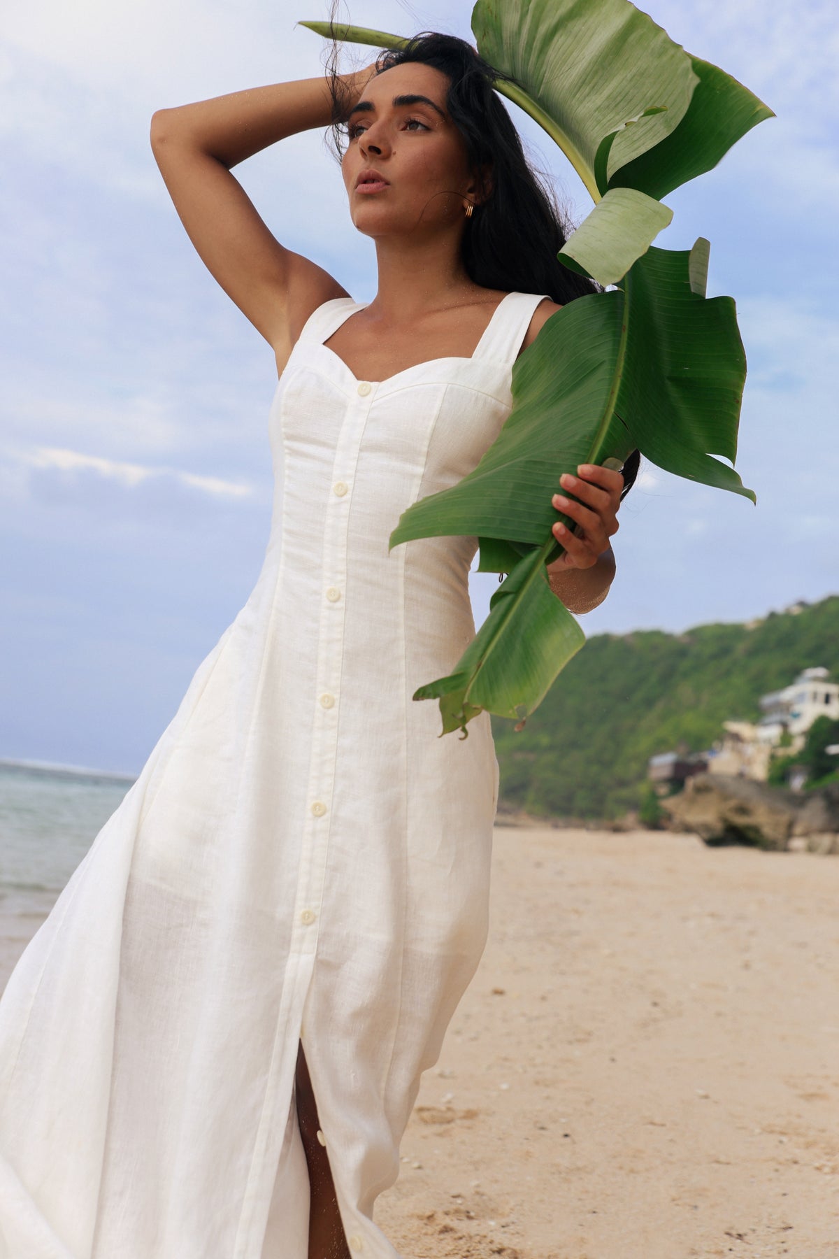 Eden Linen Midi Dress in Ivory - woman holding tropical leaf, sweetheart neckline and button-front placket, beach backdrop - Lilly Pilly Collection