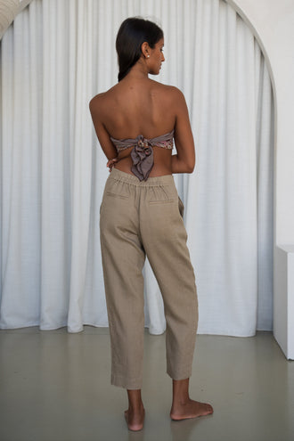 Emer Linen Pants in Taupe - back view showing elasticated waistband and tapered linen silhouette, woman wearing printed bandeau top, white curtain backdrop - Lilly Pilly Collection