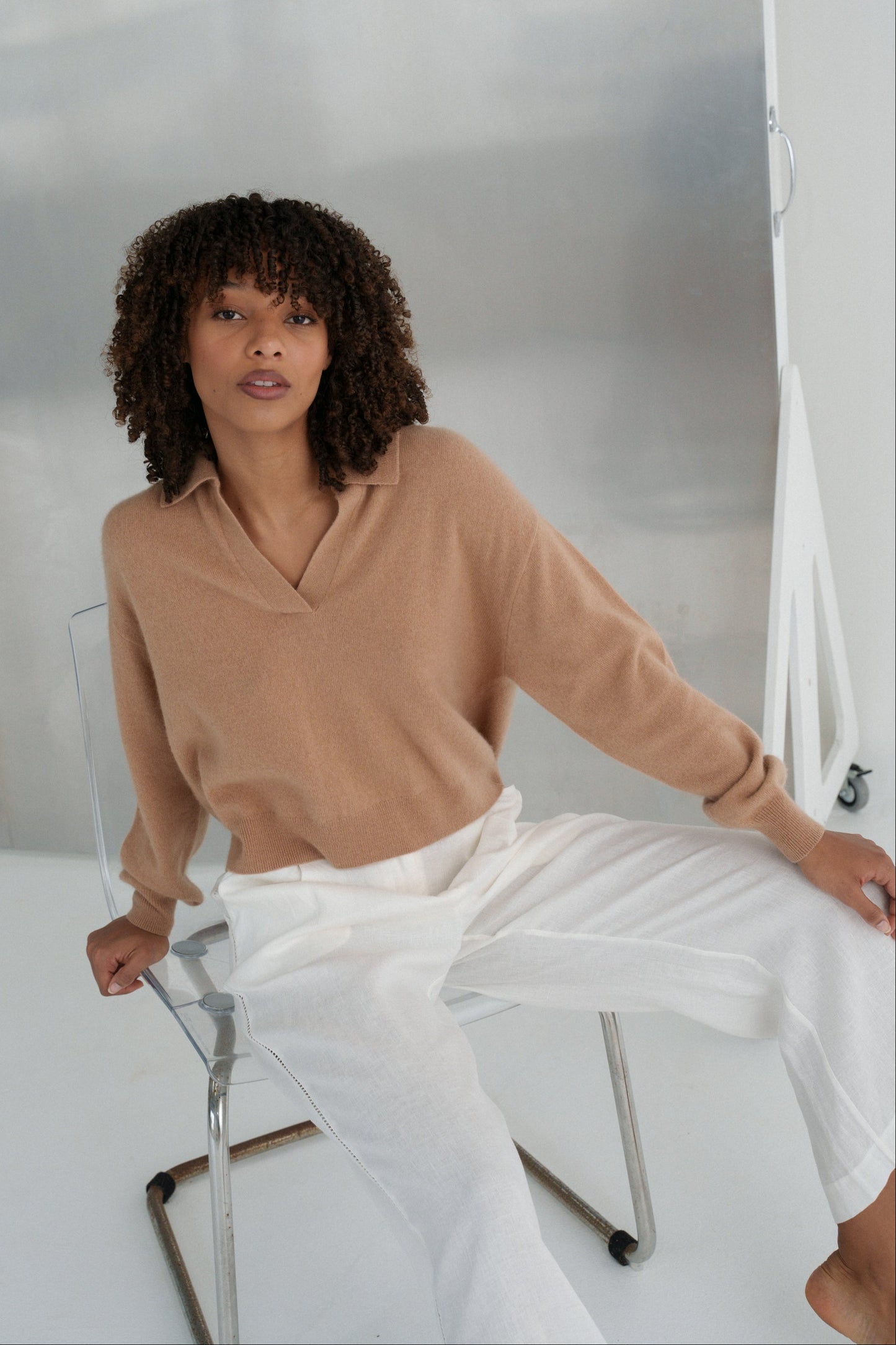 LILLY PILLY woman wearing a camel coloured Emma cashmere merino knit sweater and ivory white Ivy pants sitting on a chair in a minimalistic setting.