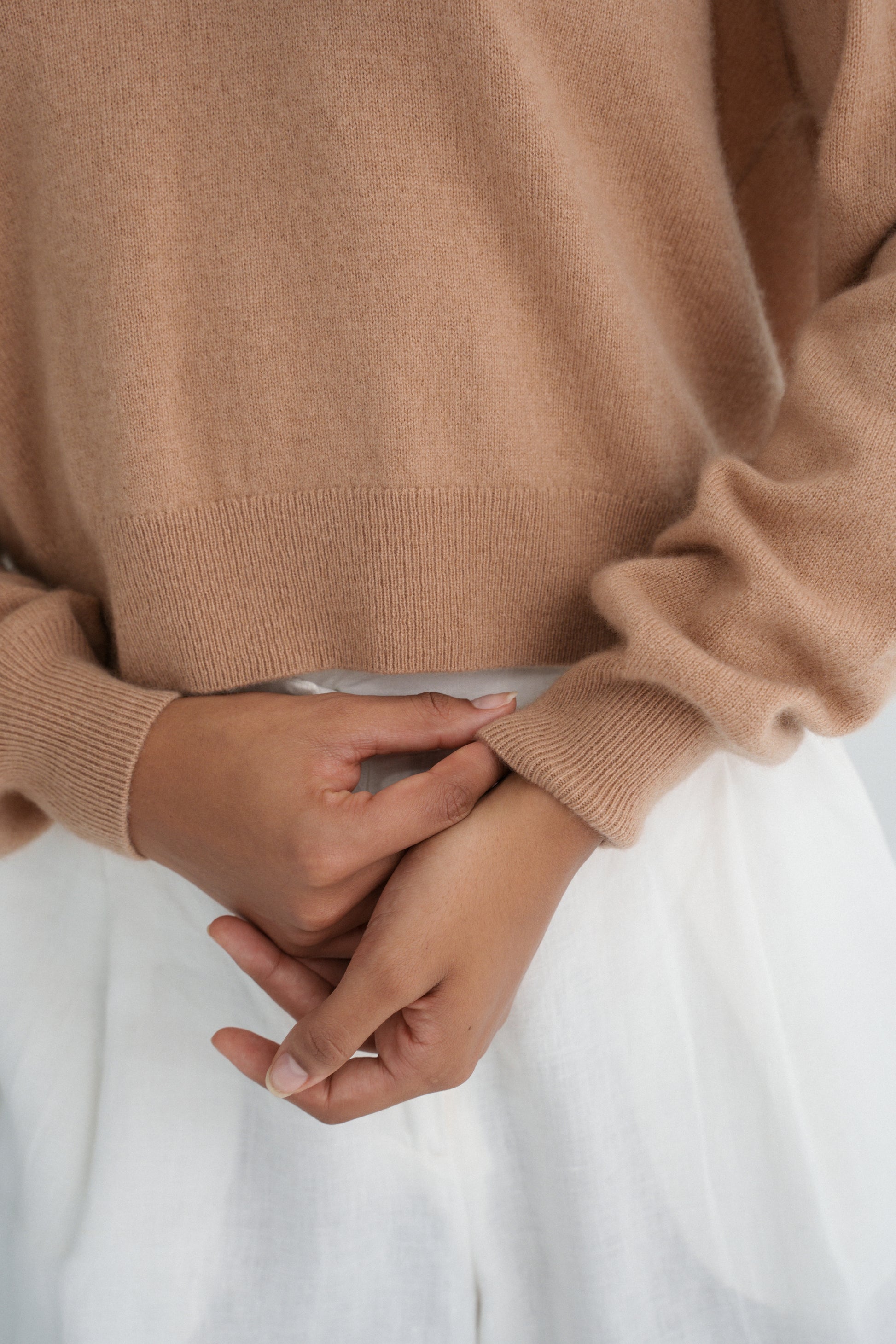 Close-up of LILLY PILLY woman wearing a camel coloured Emma cashmere merino knit sweater and ivory white Ivy pants with hands clasped together.