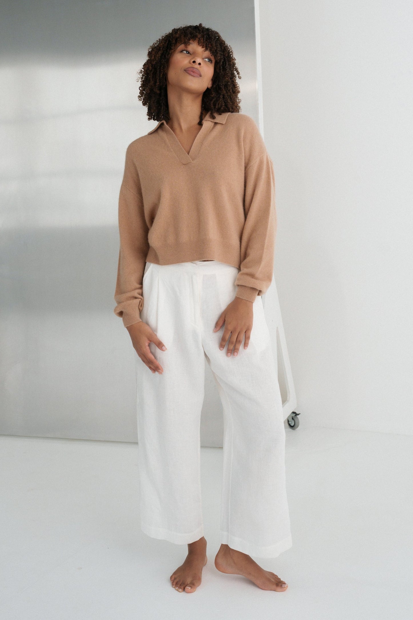LILLY PILLY woman wearing a camel coloured Emma cashmere merino knit sweater and ivory white Ivy pants standing against a plain background