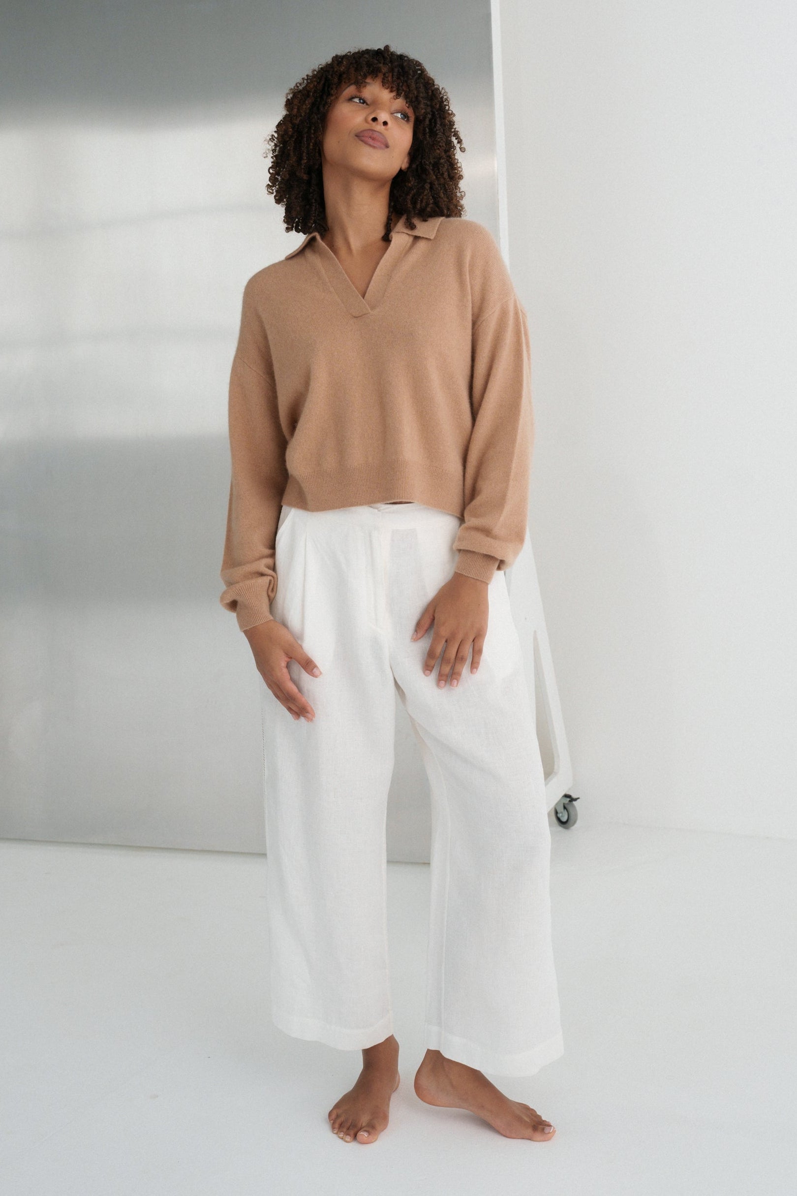LILLY PILLY woman wearing a camel coloured Emma cashmere merino knit sweater and ivory white Ivy pants standing against a plain background