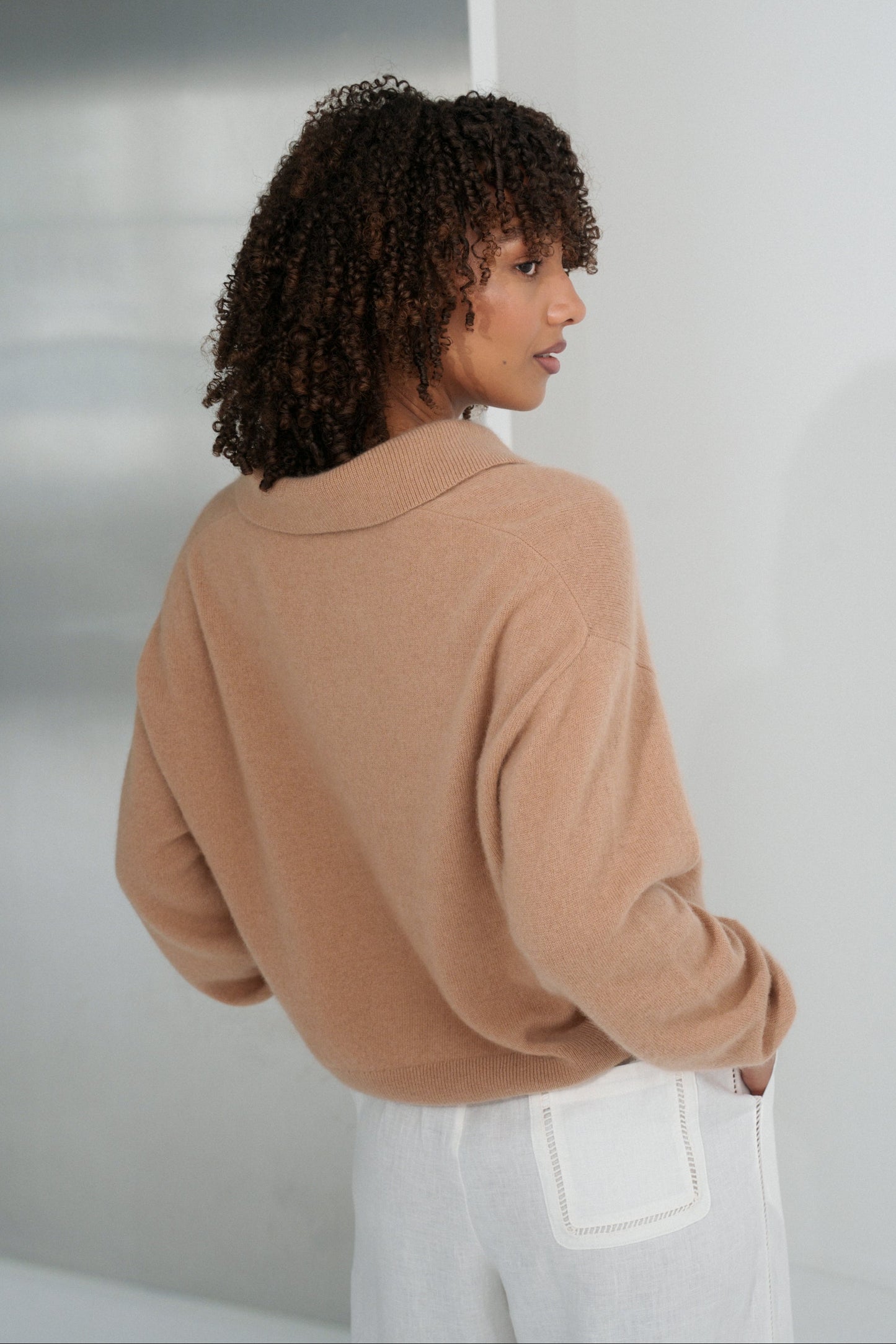 Close up back view of LILLY PILLY woman wearing a camel coloured Emma cashmere merino knit sweater and ivory white Ivy pants against a plain background