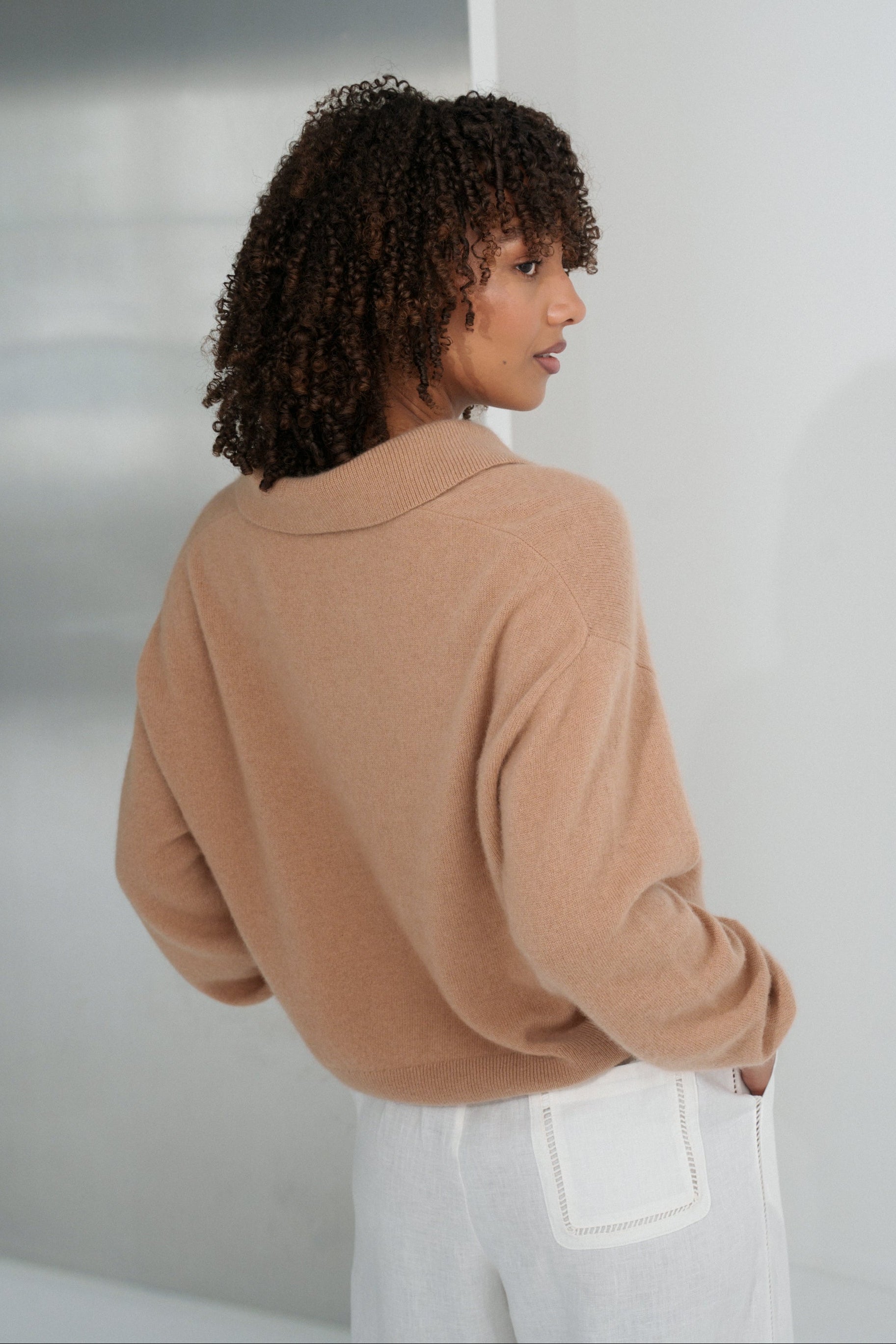 Close up back view of LILLY PILLY woman wearing a camel coloured Emma cashmere merino knit sweater and ivory white Ivy pants against a plain background