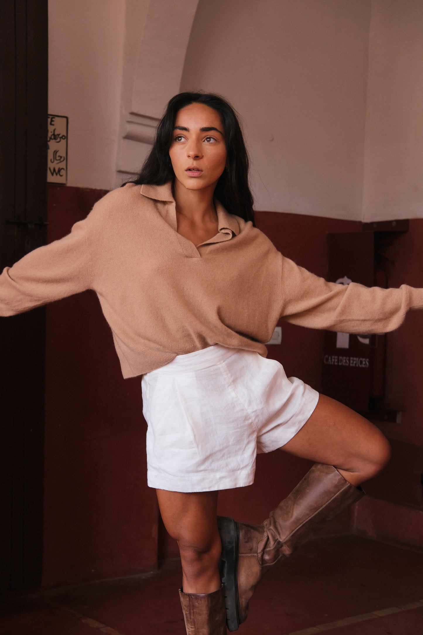 LILLY PILLY woman wearing a camel coloured Emma cashmere merino knit sweater and ivory white Gaia linen  shorts posing in a room with a red wall.