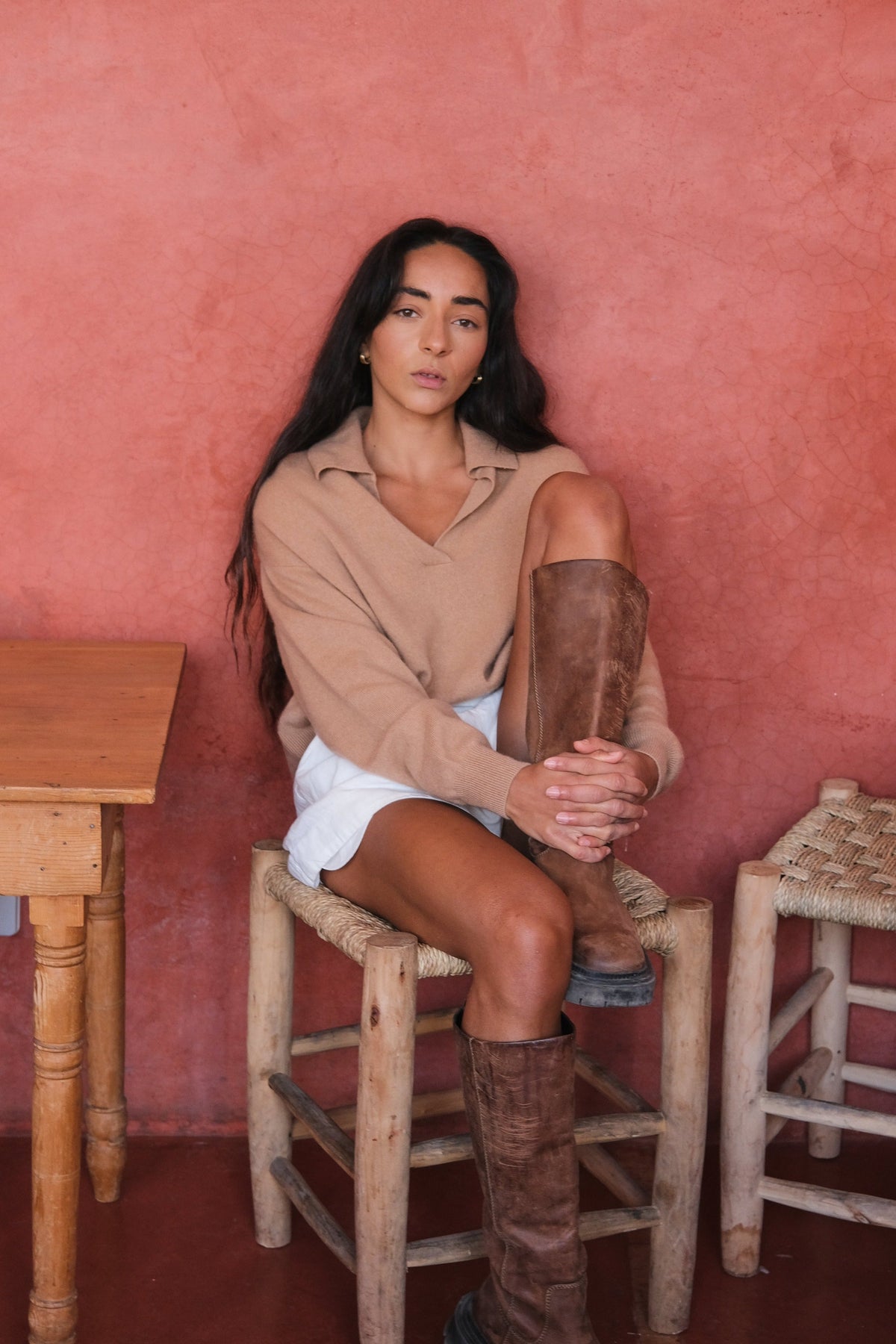 LILLY PILLY woman wearing a camel coloured Emma cashmere merino knit sweater and ivory white Gaia linen  shorts sitting on a wooden stool against a pink wall with a wooden table and chair in the foreground.