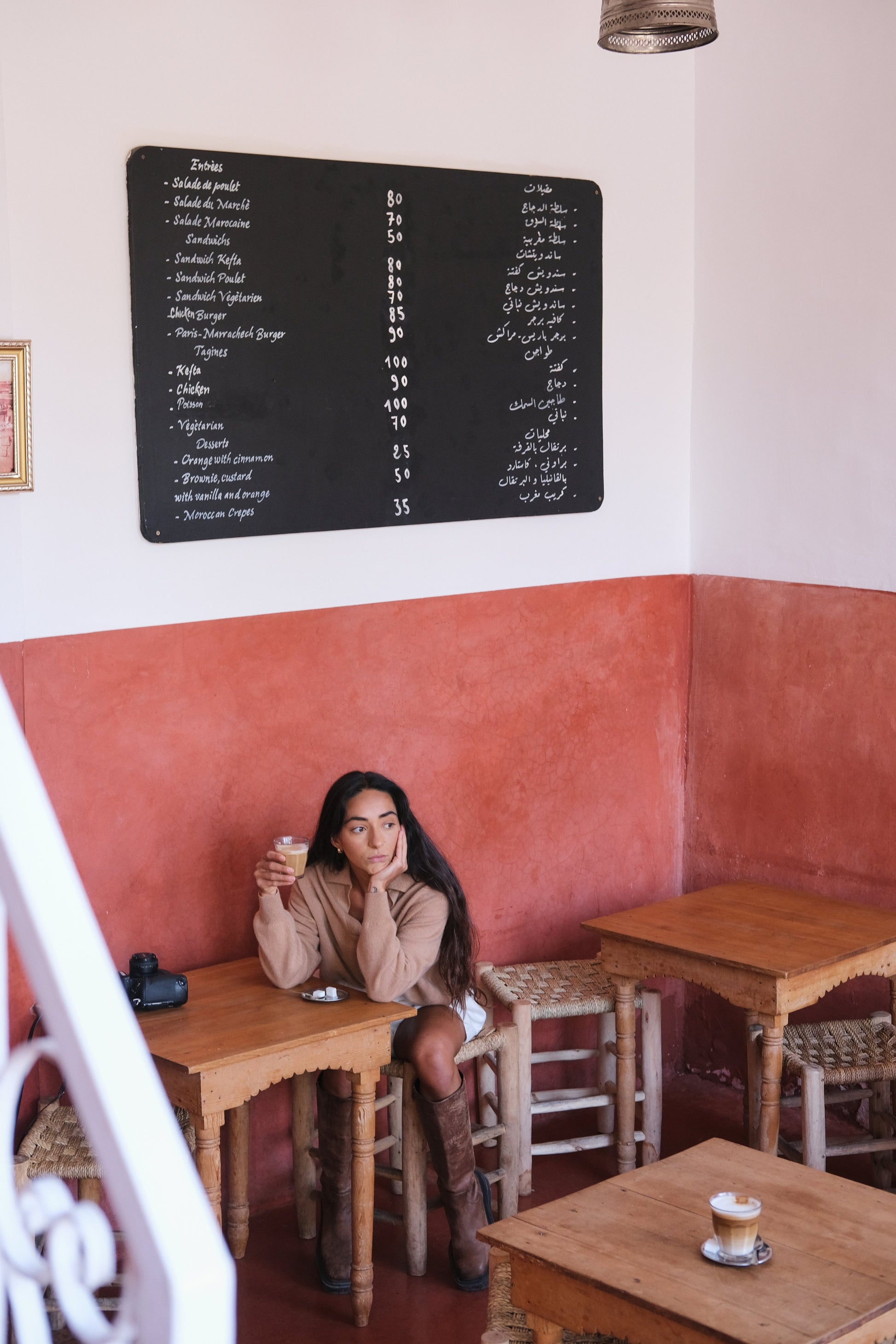 LILLY PILLY woman wearing a camel coloured Emma cashmere merino knit sweater and ivory white Gaia linen  shorts sitting at a table in a cafe with a menu board on the wall.