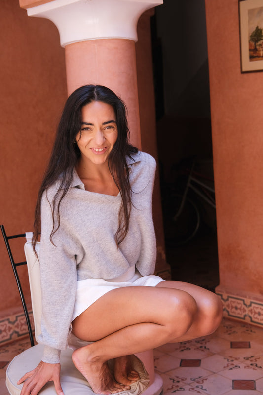 LILLY PILLY woman wearing a grey marle coloured Emma cashmere merino knit sitting on a chair in a warm-toned room with a decorative column.
