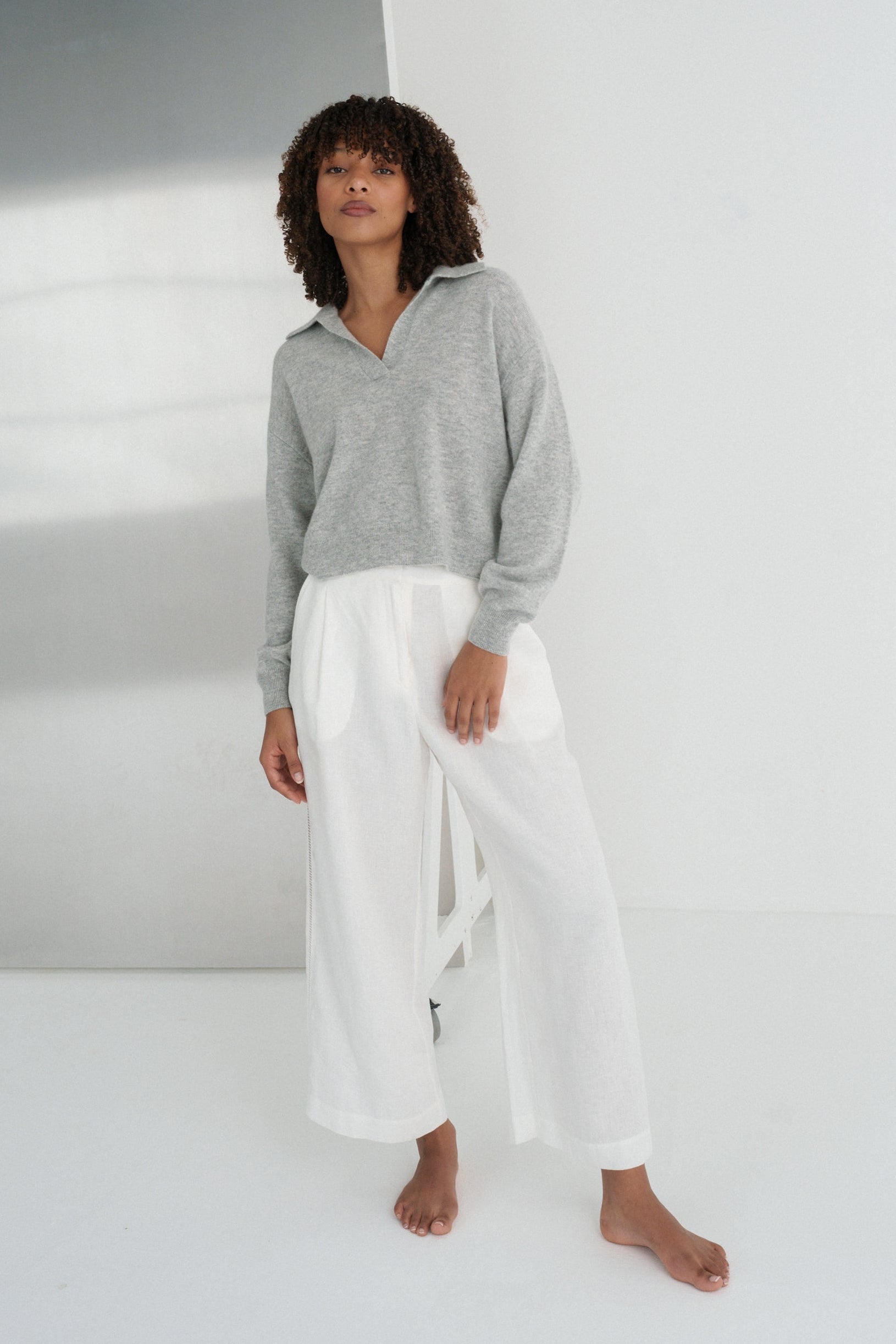 LILLY PILLY woman wearing a grey marle coloured Emma cashmere merino knit wearing a grey sweater and white pants standing against a white wall with a reflective surface.