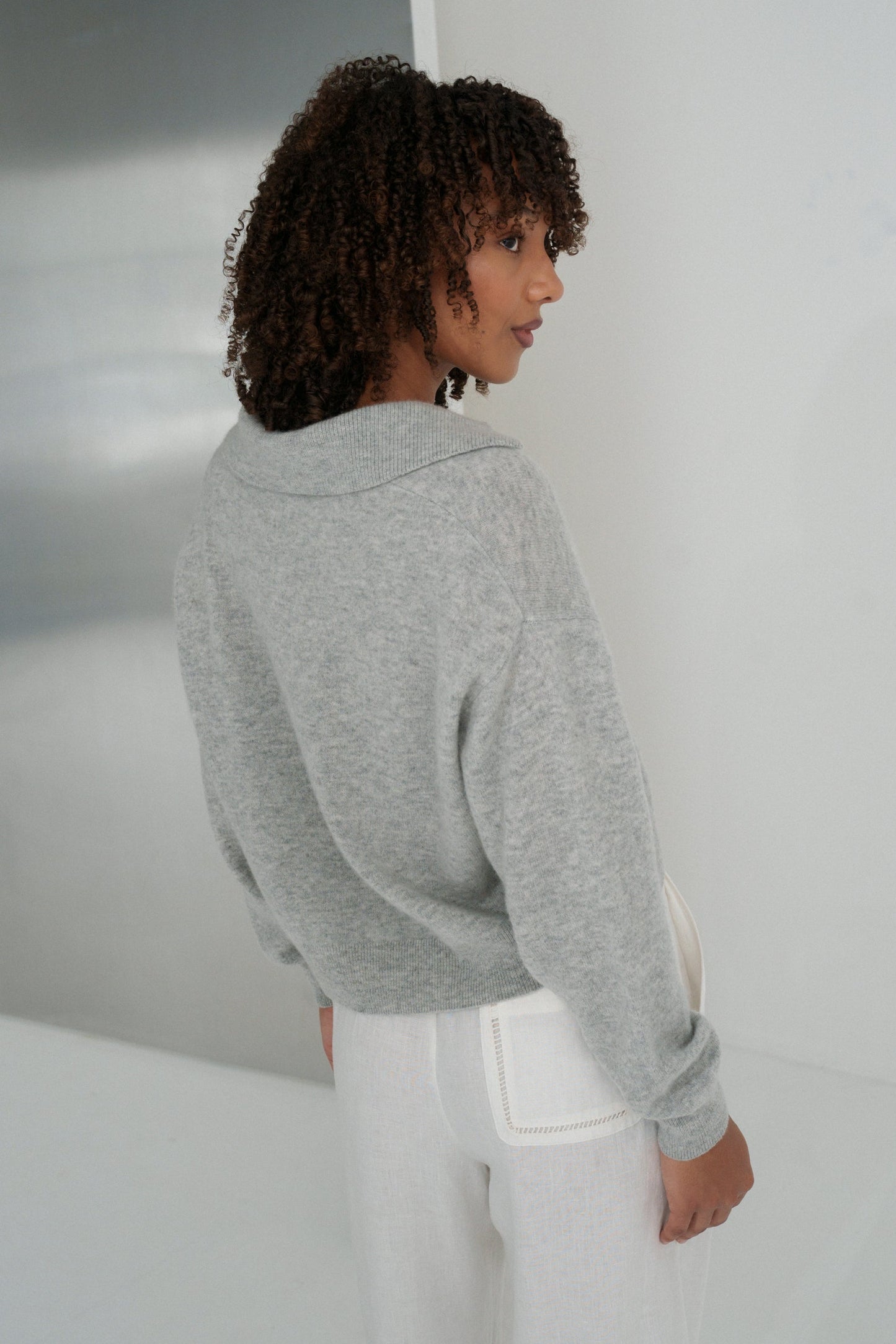 Back view of LILLY PILLY woman wearing a grey marle coloured Emma cashmere merino knit sweater and ivory white pants in a minimalistic setting