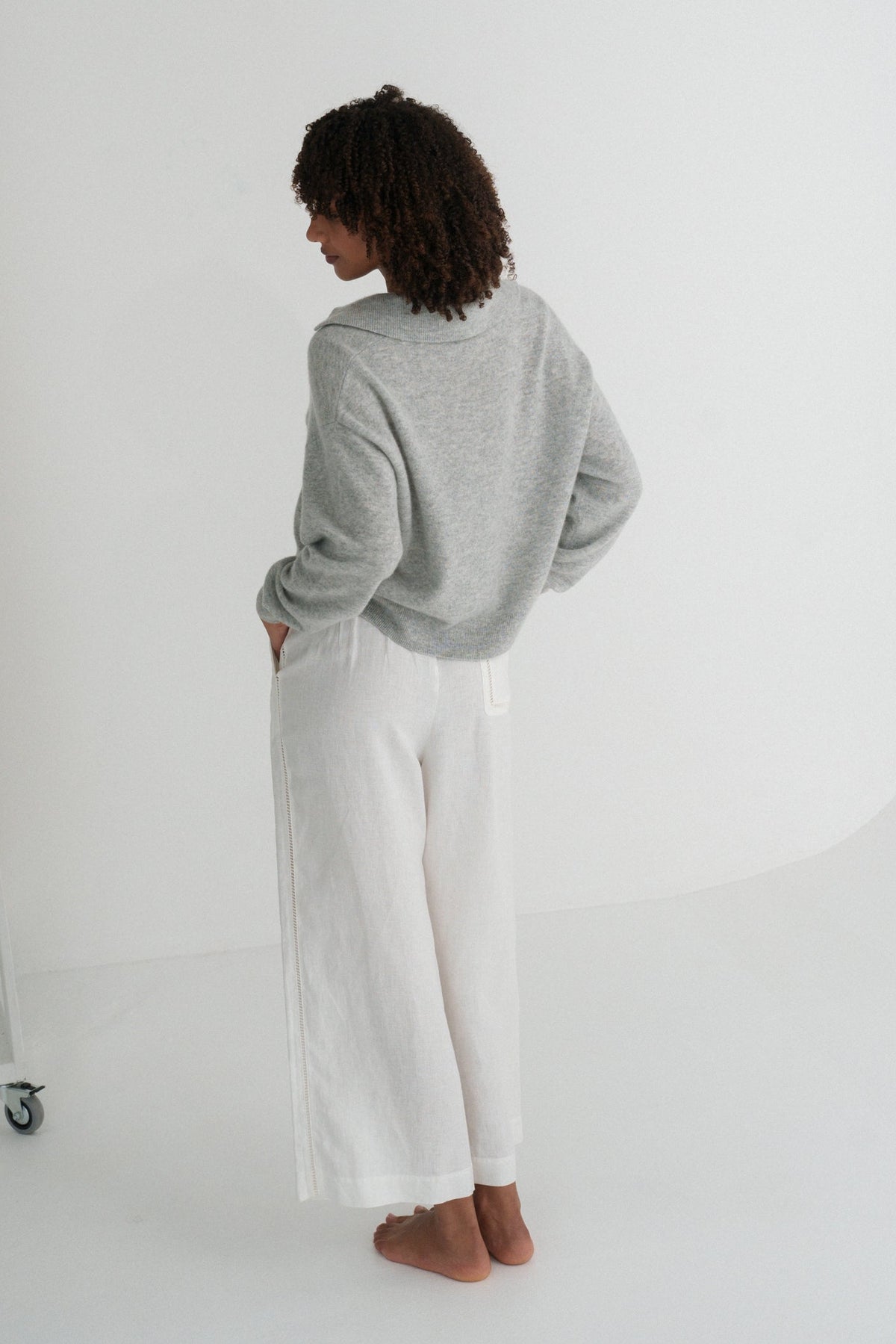 Back view of LILLY PILLY woman wearing a grey marle coloured Emma cashmere merino knit sweater and ivory white pants standing in a minimalistic room.