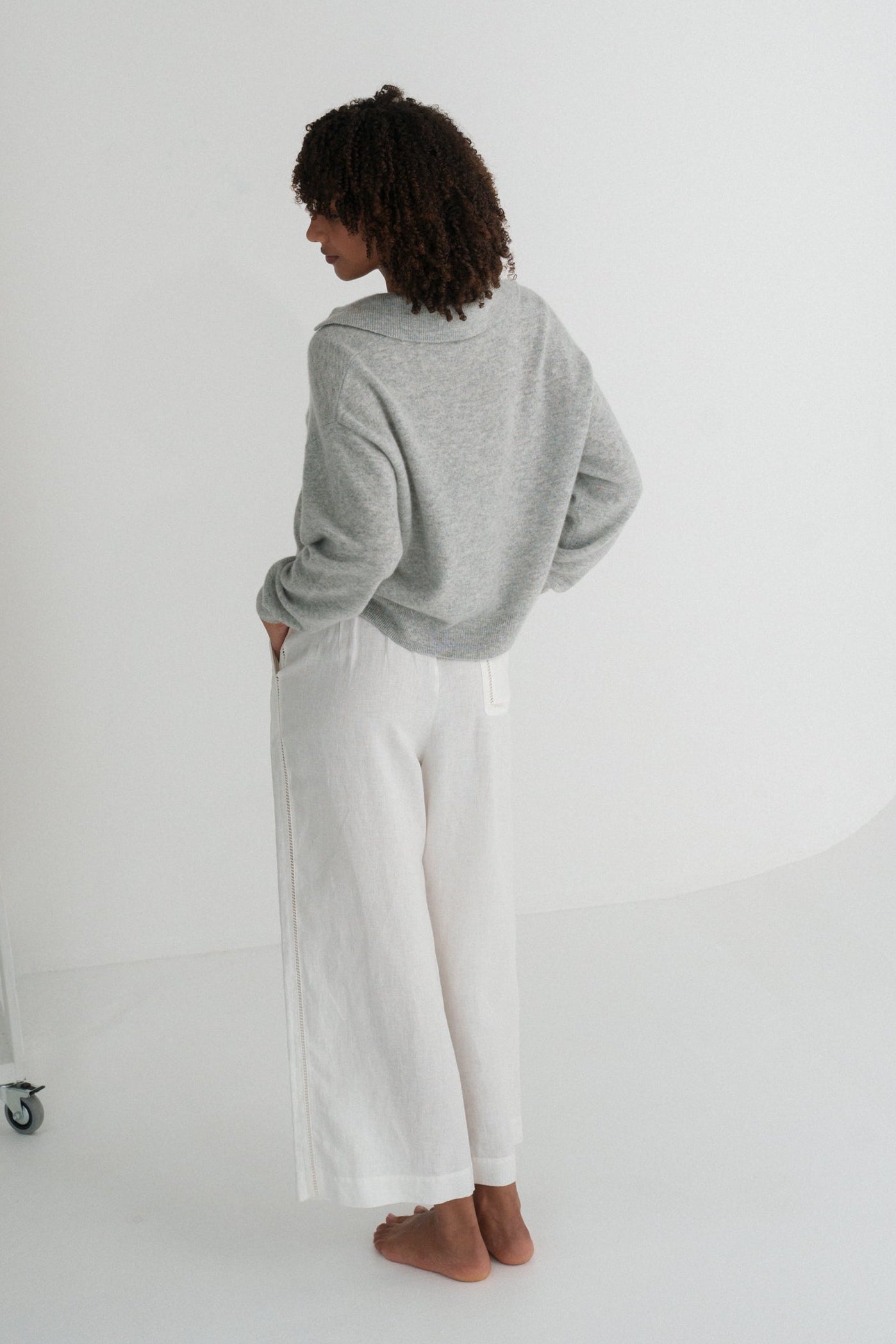 Back view of LILLY PILLY woman wearing a grey marle coloured Emma cashmere merino knit sweater and ivory white pants standing in a minimalistic room.