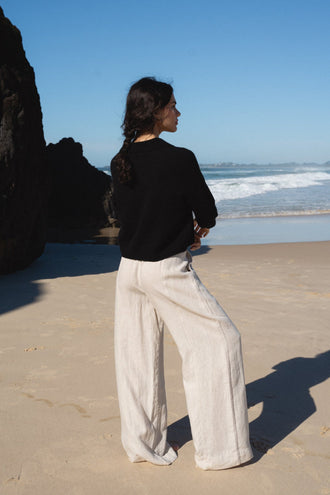 Emma Cashmere Knit in Black - back view, standing on beach with ocean and rocks backdrop, styled with Oli Linen Pants in Oatmeal - Lilly Pilly Collection