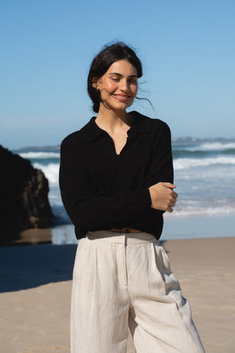 Emma Cashmere Knit in Black - close-up front view, smiling, arms crossed, beach backdrop, styled with Oli Linen Pants in Oatmeal - Lilly Pilly Collection