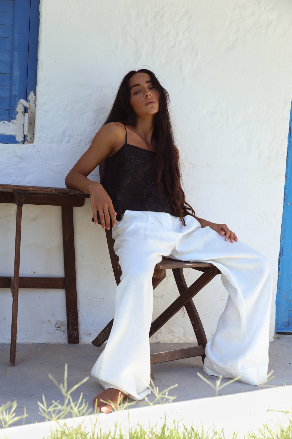 Flora Ramie Cami in Black - seated, floral embroidery and thin straps, worn with Oli Pants in Ivory, blue shutters backdrop - Lilly Pilly Collection