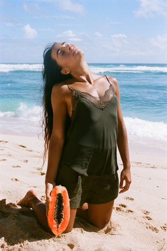 Frankie Silk Cami in Khaki - woman kneeling on sand, V-neckline and lace trim, styled with matching linen shorts, holding papaya, beach - Lilly Pilly Collection