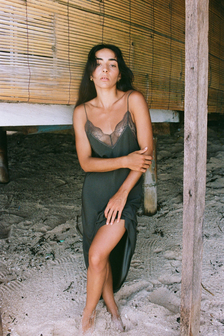 Freya Khaki Silk Slip Dress - woman leaning against rustic wooden post, lace V-neckline detail in khaki silk - Lilly Pilly Collection
