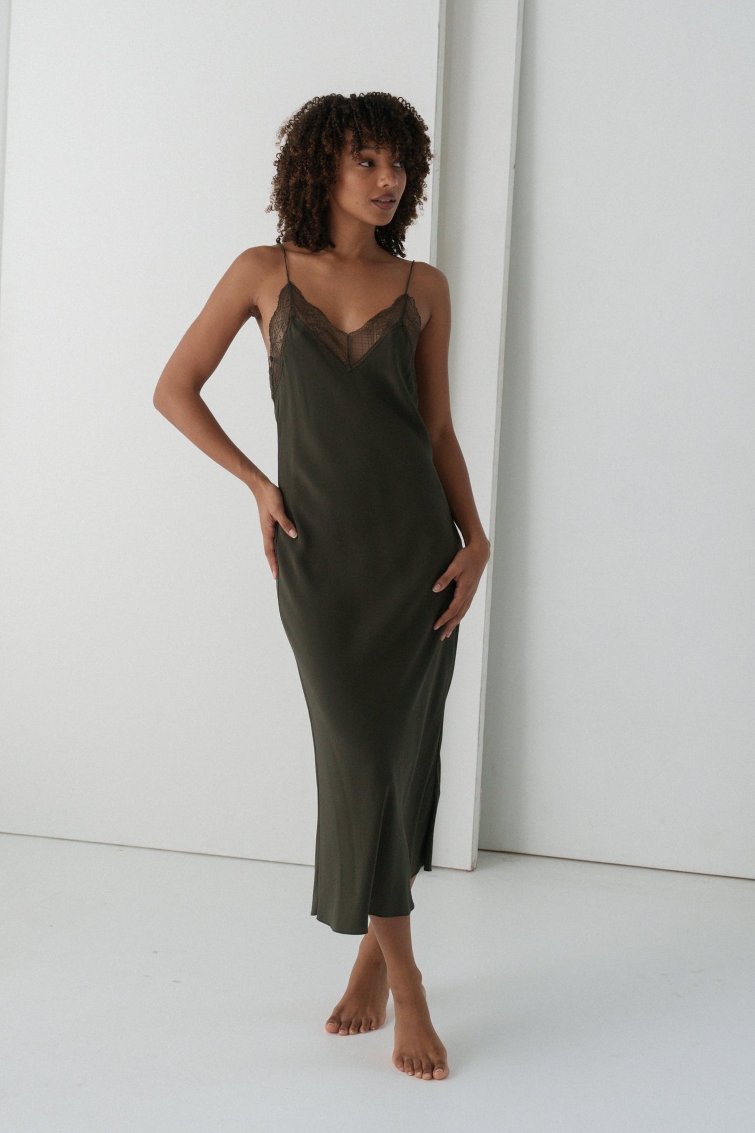 Woman wearing a khaki Freya silk slip dress standing in a minimal white room.