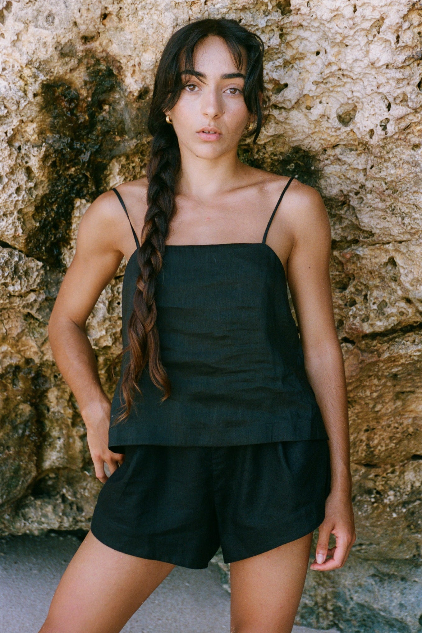 Woman wearing a black linen silk tank top and Gaia linen shorts against a stone wall.