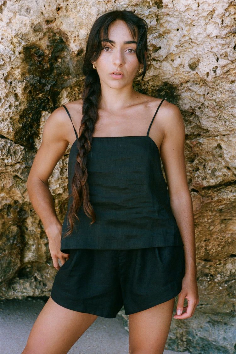 Gaia Linen Shorts in Black - full length front view, woman wearing Black linen shorts with matching black linen square-neck cami, coastal stone wall backdrop - Lilly Pilly Collection
