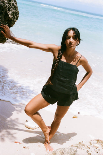 Gaia Linen Shorts in Black - three-quarter front view, woman wearing Black linen shorts with matching black linen cami, leaning on coastal rock, sandy beach and ocean backdrop - Lilly Pilly Collection