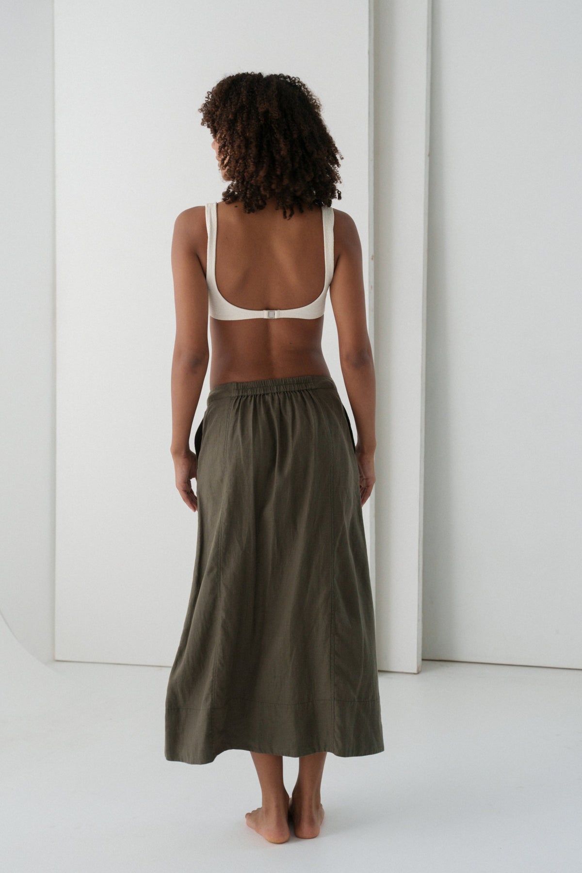 Back view of woman wearing a white tank top and khaki green Gia linen skirt against a white background