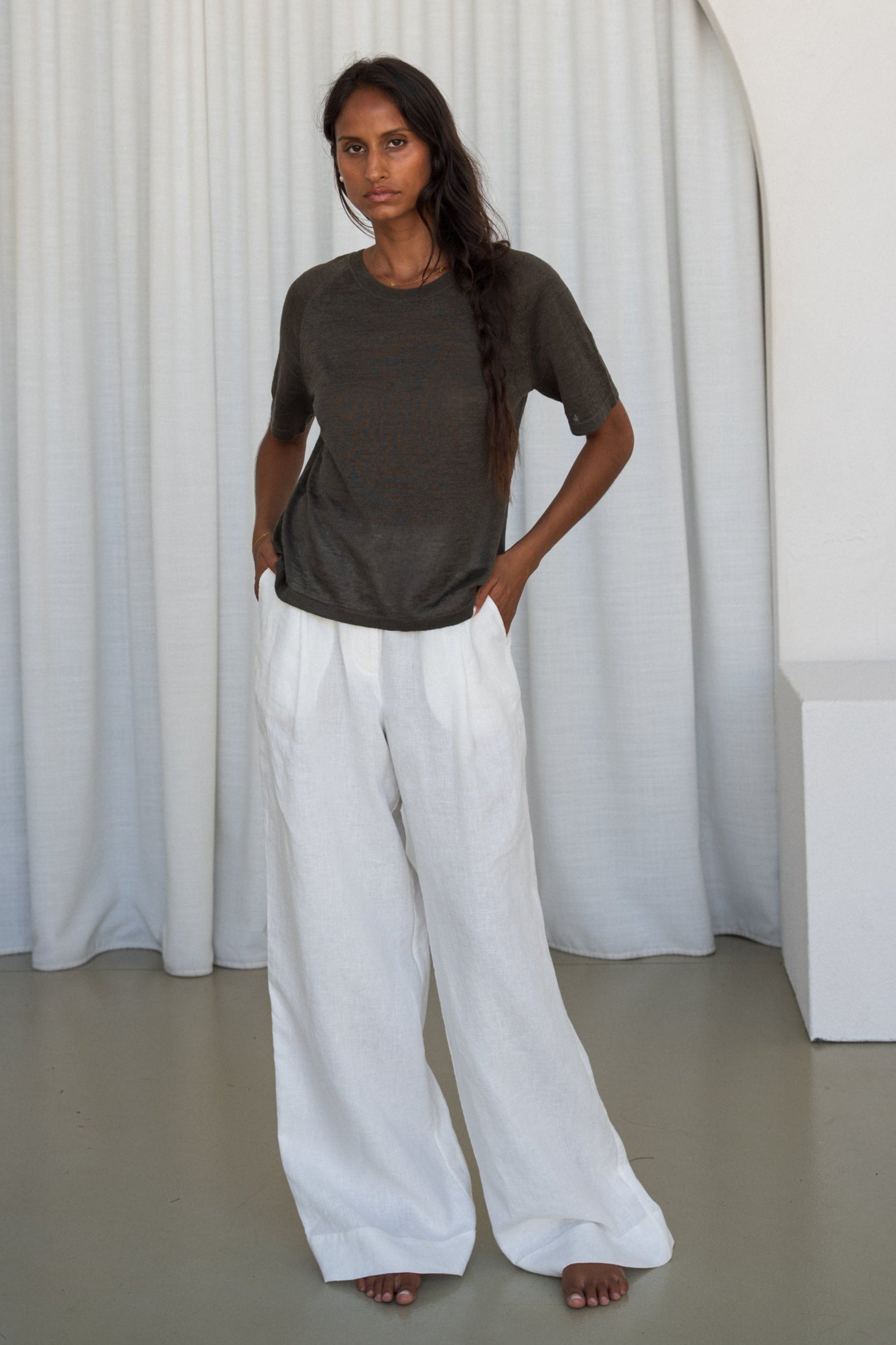 Woman wearing a khaki Grace linen knit top and Oli ivory linen pants with white curtain wall background
