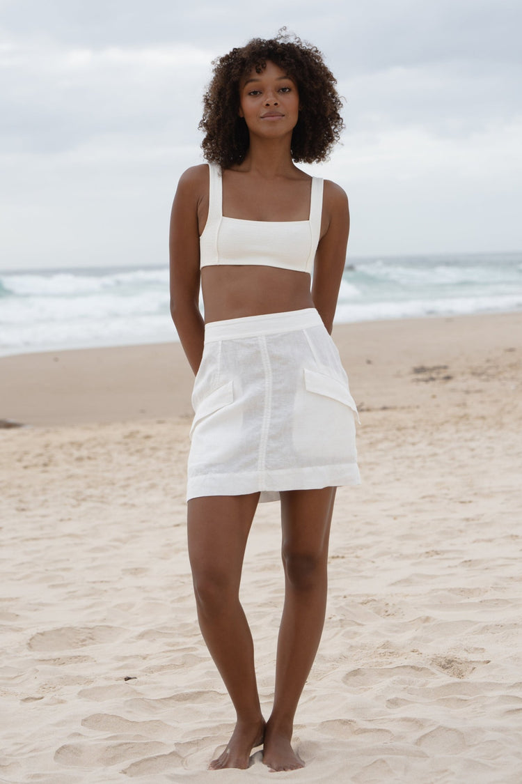 Ilaria Linen Mini Skirt in Ivory - full length front view, cargo pocket detail, worn with square-neck bralette, standing on beach with ocean backdrop - Lilly Pilly Collection