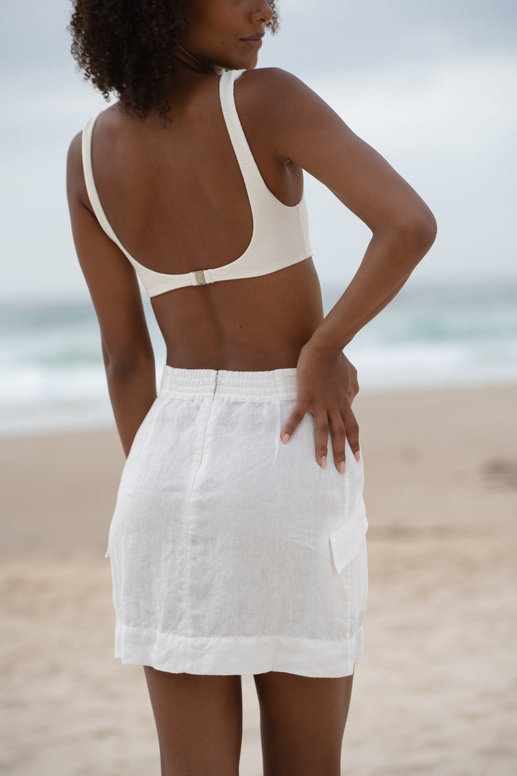 Ilaria Linen Mini Skirt in Ivory - close-up back view, elasticated waistband, worn with square-neck bralette, standing on beach - Lilly Pilly Collection