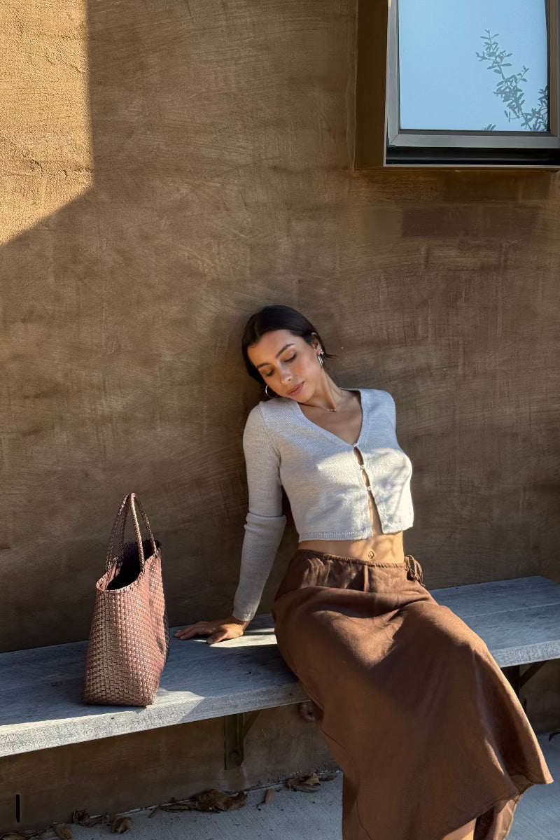 LILLY PILLY woman sitting on a bench against a textured wall wearing an Indie merino wool knit top in grey, with Riley chocolate linen skirt  and a woven handbag beside her.