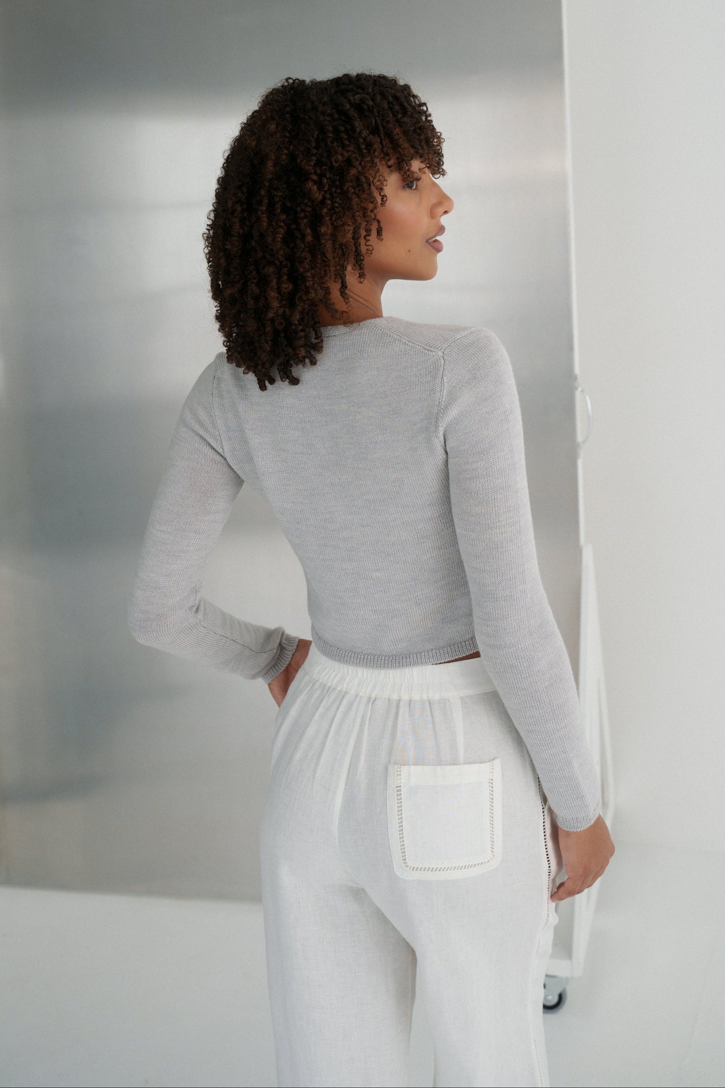 back view LILLY PILLY woman wearing a fog gray Indie merino wool cardigan top and ivory white pants in a minimalistic setting.