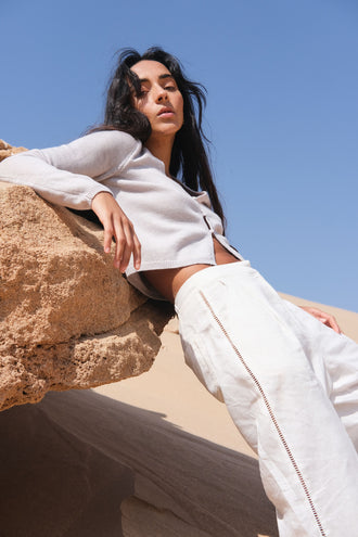 Indie Merino Cardi Knit in Fog Grey - front view, styled with ivory Rika pants, seated on a rock against a clear blue sky - Lilly Pilly Collection