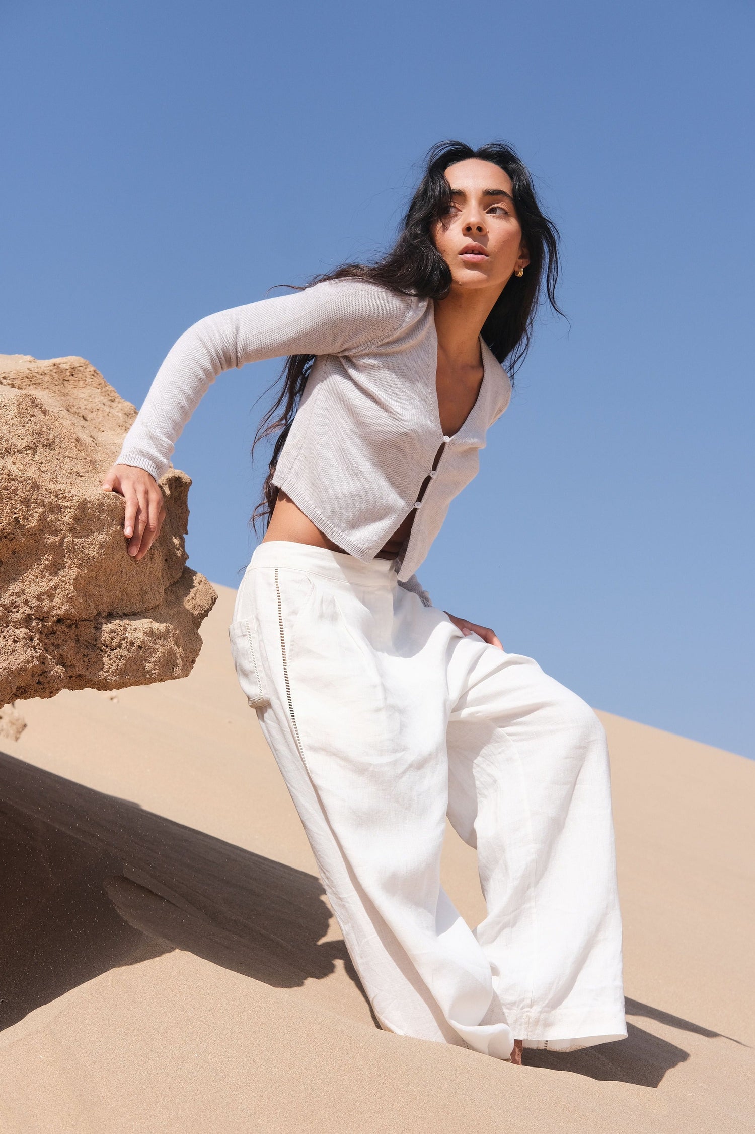 LILLY PILLY woman wearing a fog gray Indie merino wool cardigan and ivory Rika pants standing on sand with a clear blue sky