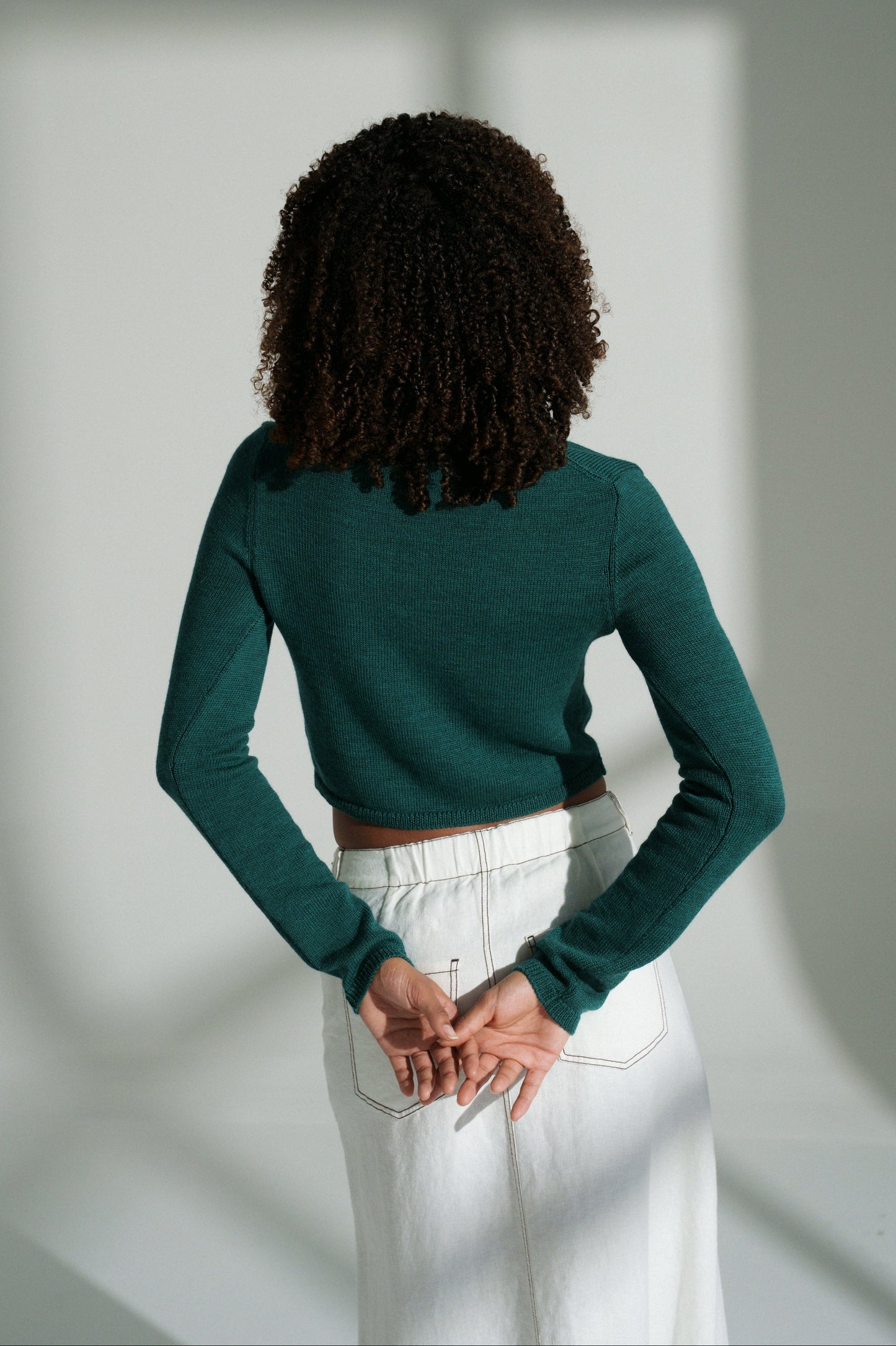 Back view of LILLY PILLY woman wearing a pine green Indie merino wool cardigan and white skirt standing in front of a window with light filtering through.