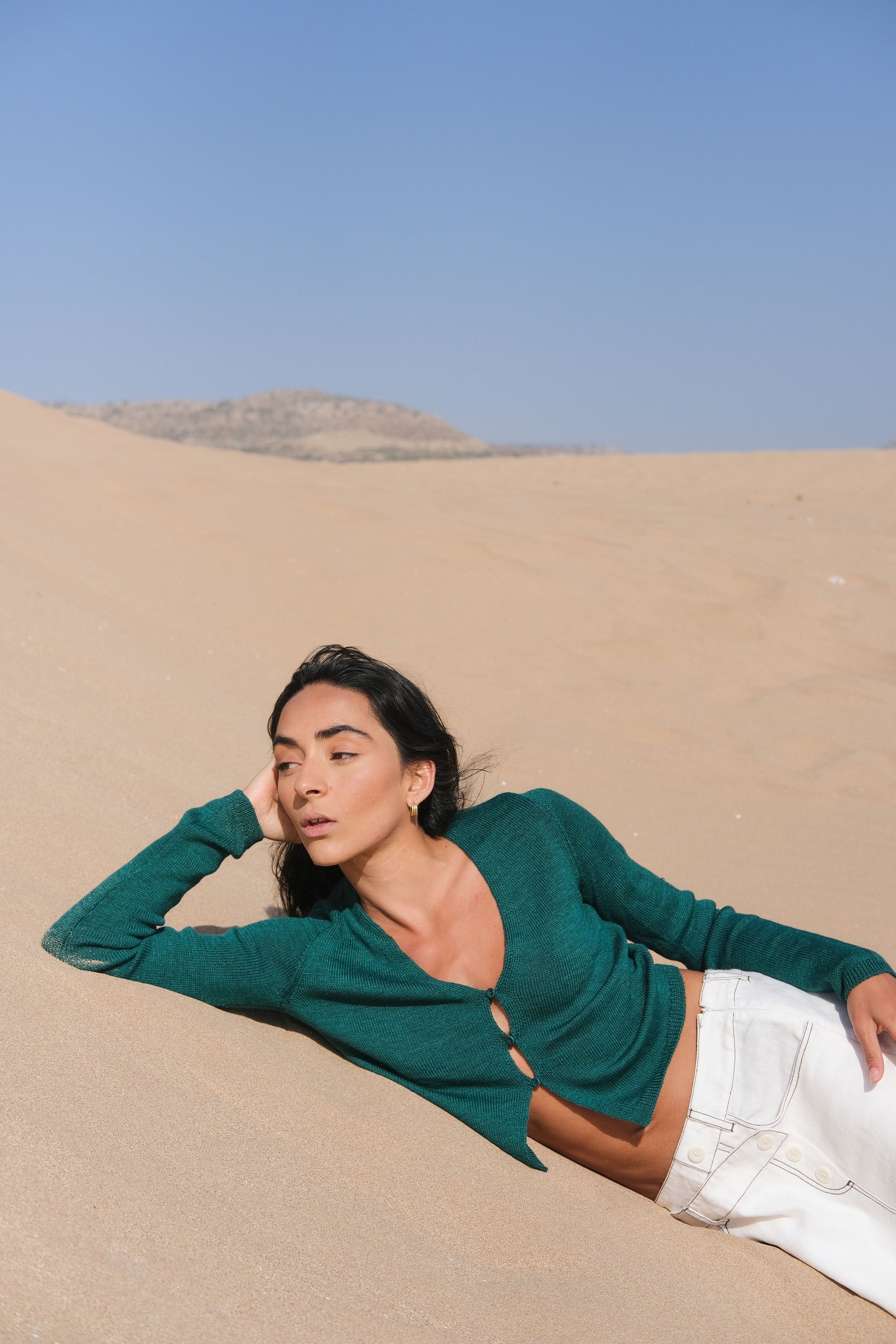 LILLY PILLY woman wearing a pine green Indie merino wool cardigan and ivory white skirt lying on sand dunes wearing a green top and white pants with a clear blue sky.