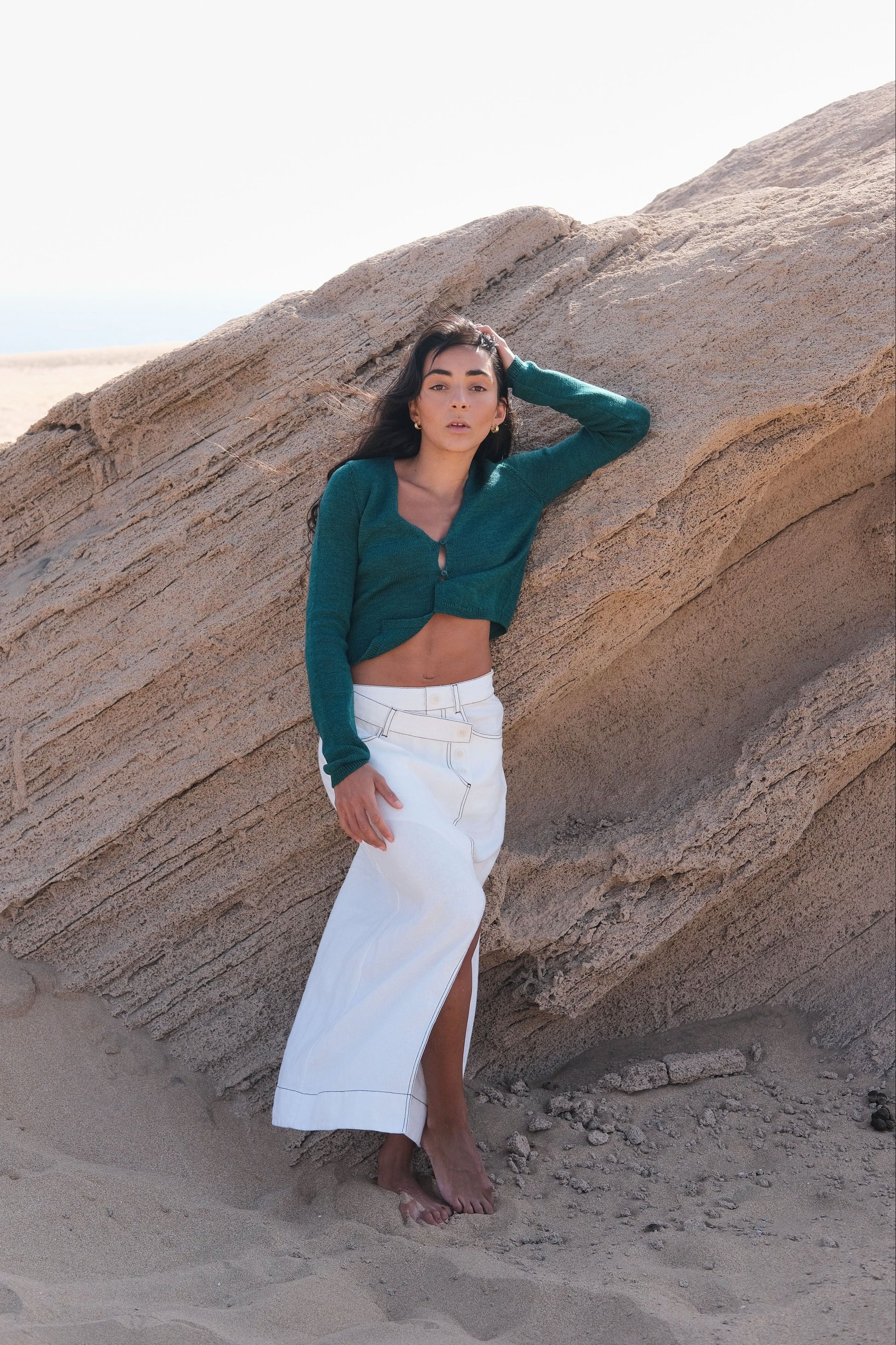 LILLY PILLY woman wearing a pine green Indie merino wool cardigan and white skirt standing against a large rock formation on a sandy beach.
