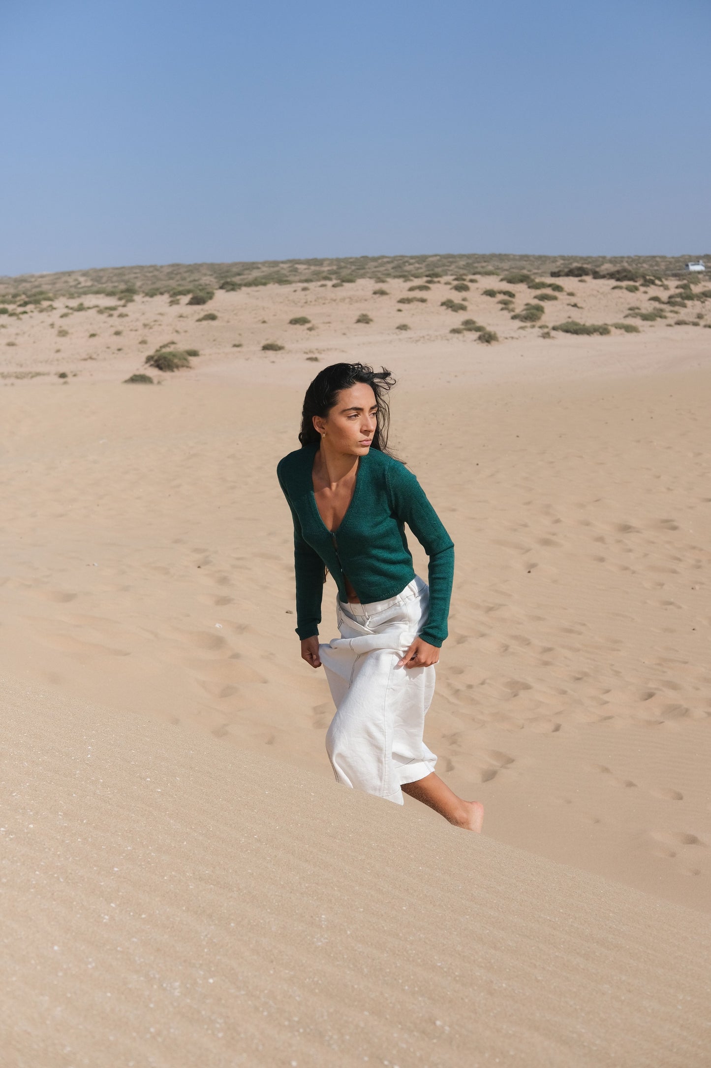 LILLY PILLY woman wearing a pine green Indie merino wool cardigan and white skirt standing in a desert landscape with clear blue sky.