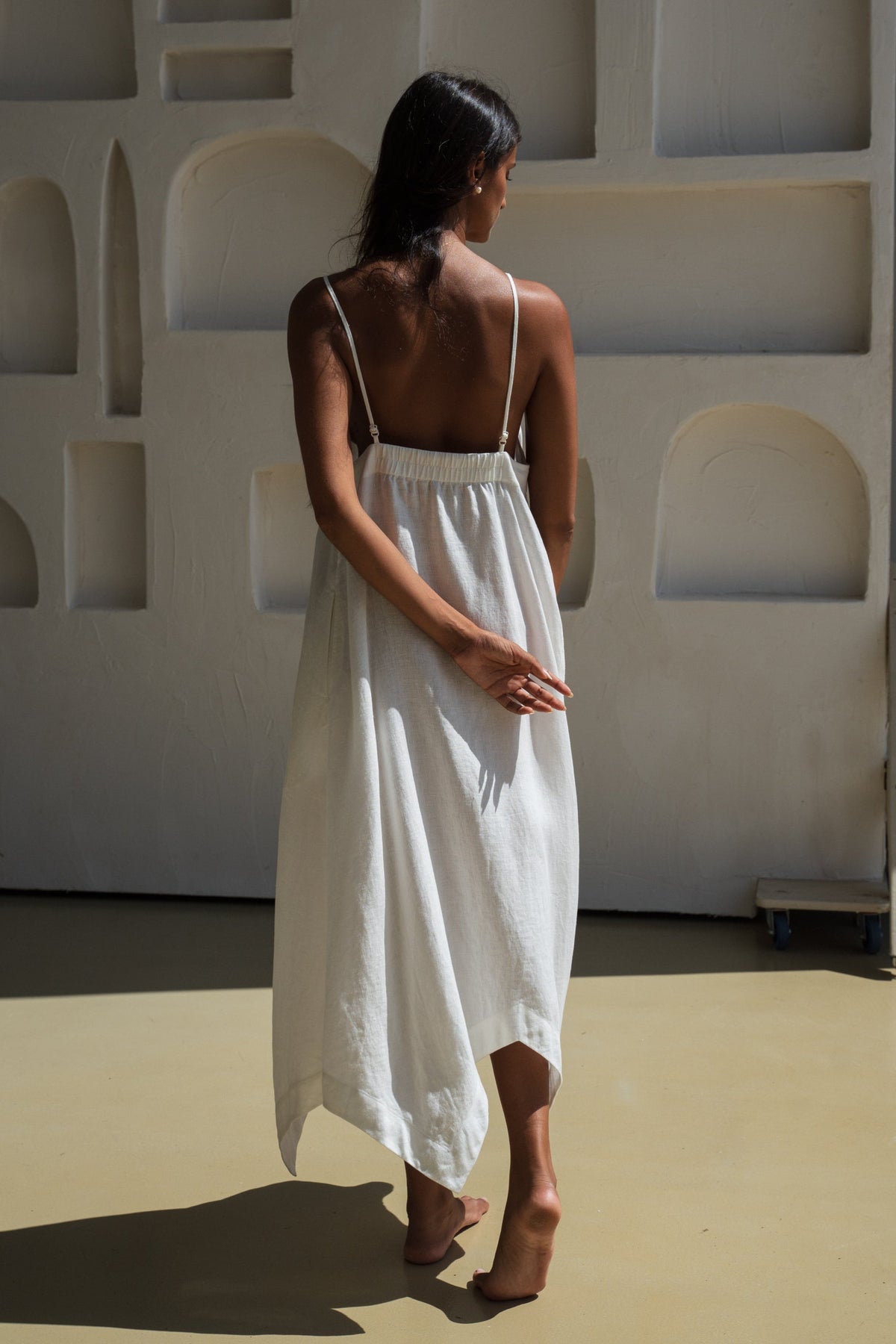 Back view of a woman wearing a ivory white Inez linen dress standing against a minimalist wall with a textured wall.