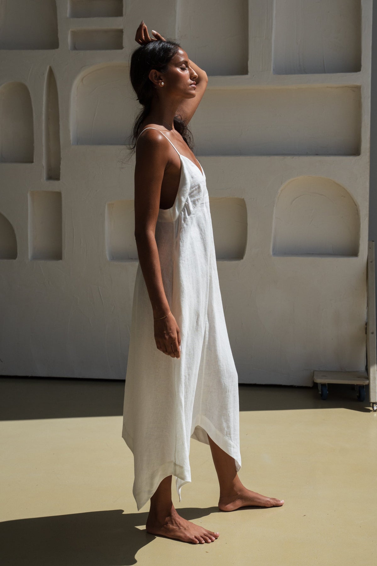 Side view of a woman wearing a ivory white Inez linen dress standing against a minimalist wall with geometric shapes. w