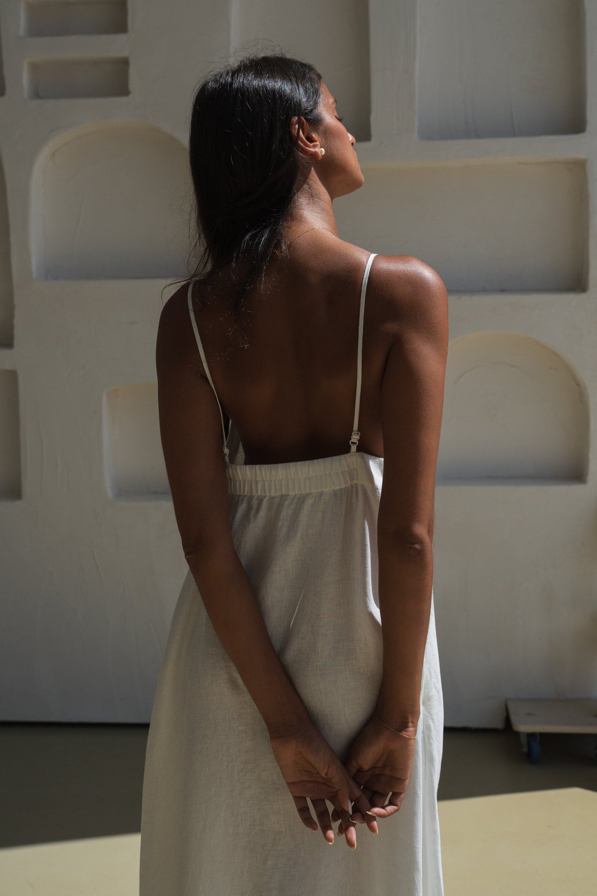 Close up back view of a woman wearing a ivory white Inez linen dress standing against a minimalistic wall.