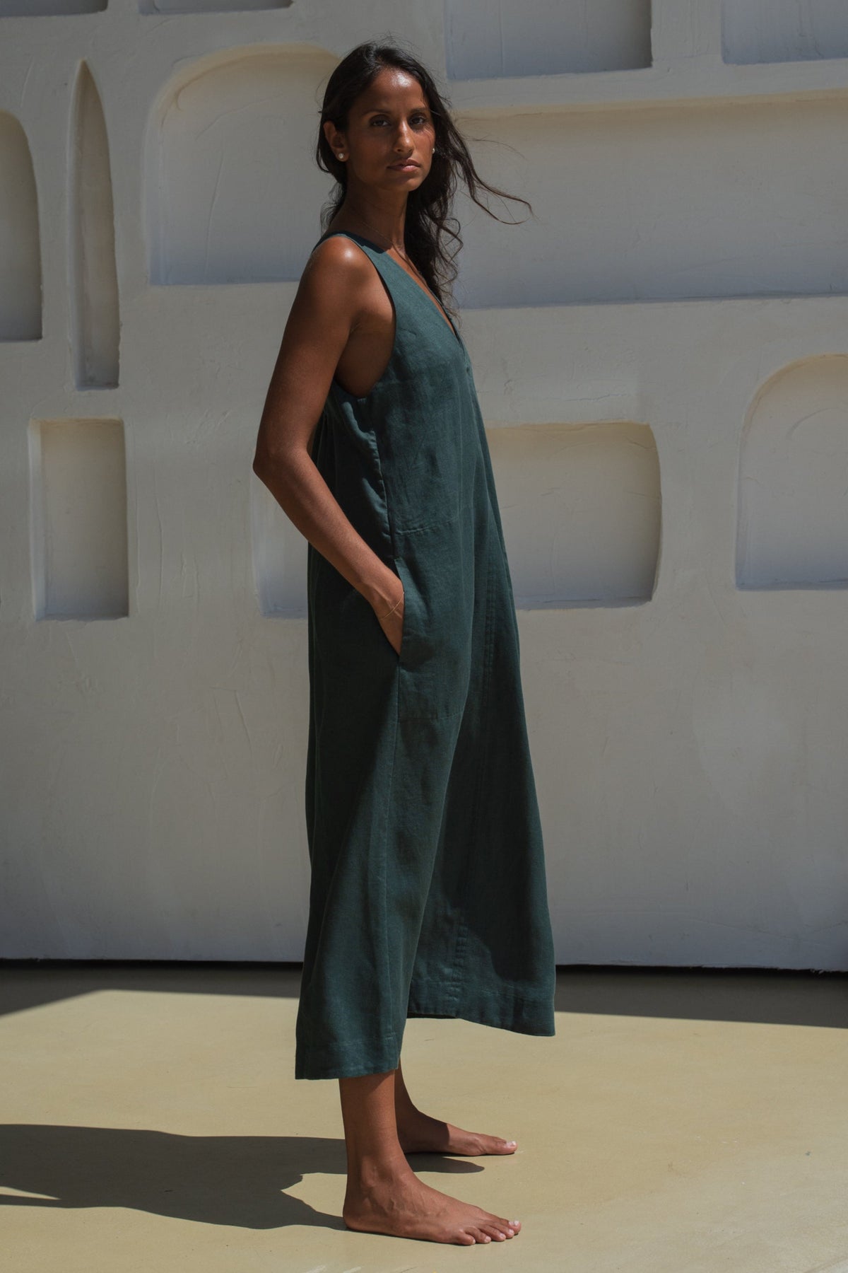 Woman in an forest green LILLY PILLY Iris dress showing the side view standing in front of a white wall with geometric shapes.