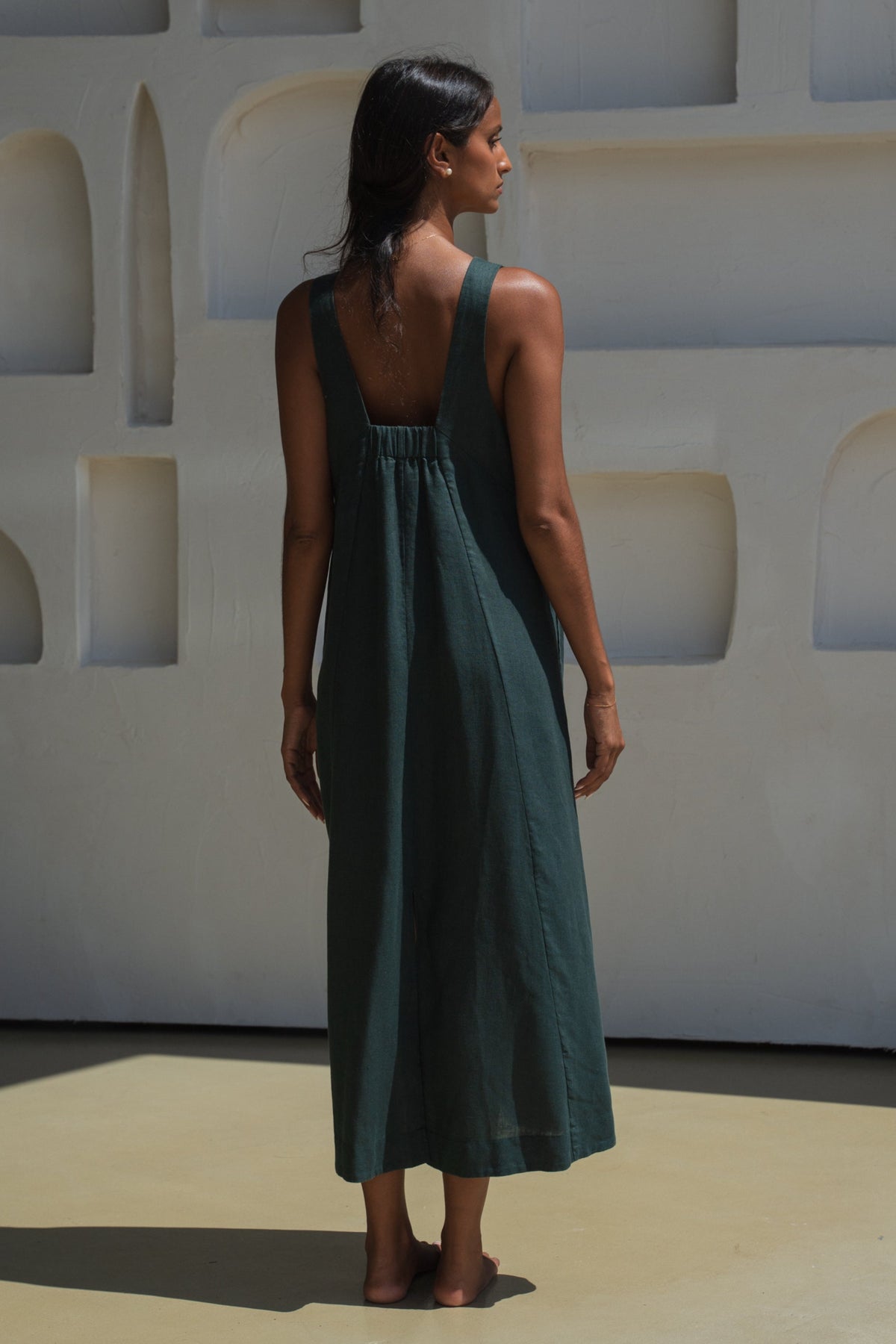 Woman in an forest green LILLY PILLY Iris dress showing the back design view standing in front of a white wall with geometric shapes.