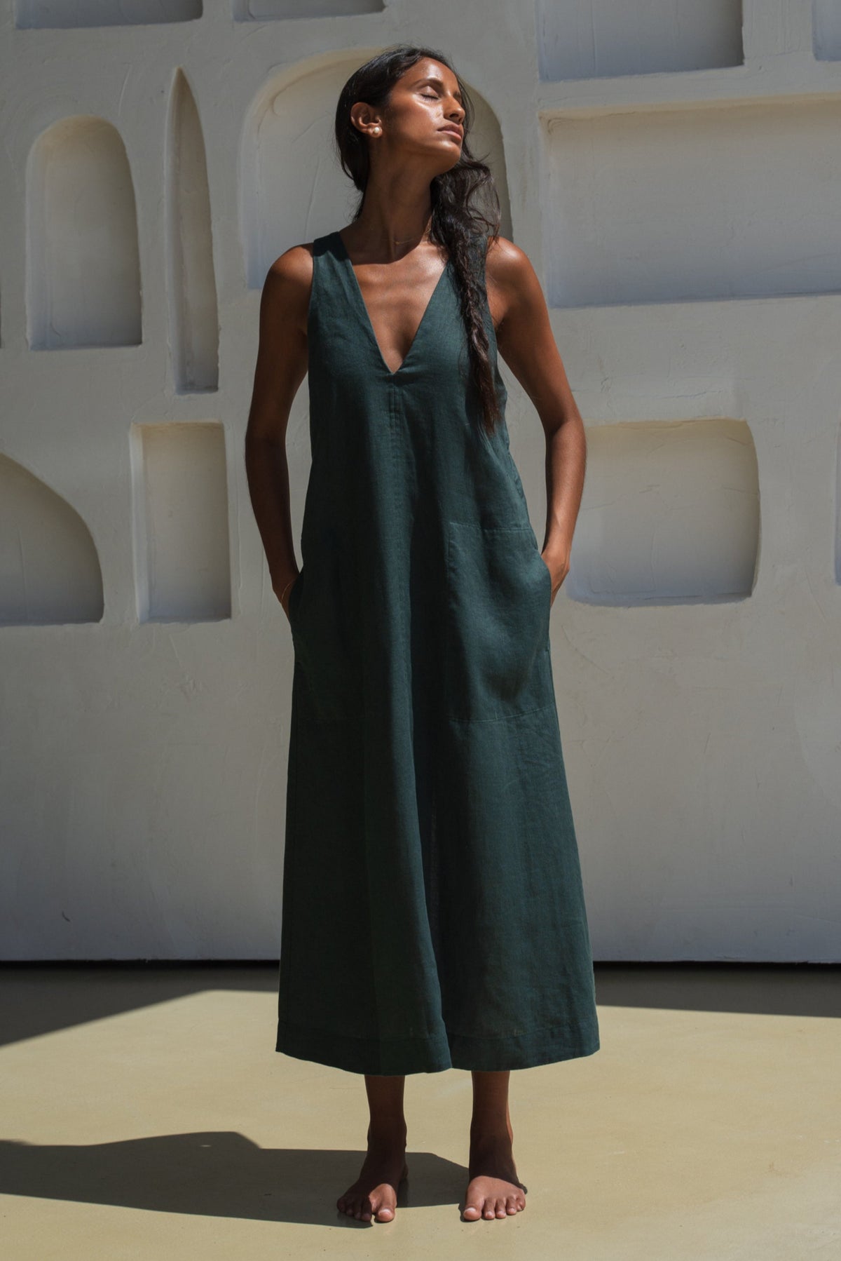 Woman in a forest green LILLY PILLY Iris dress standing against a white wall with geometric shapes.