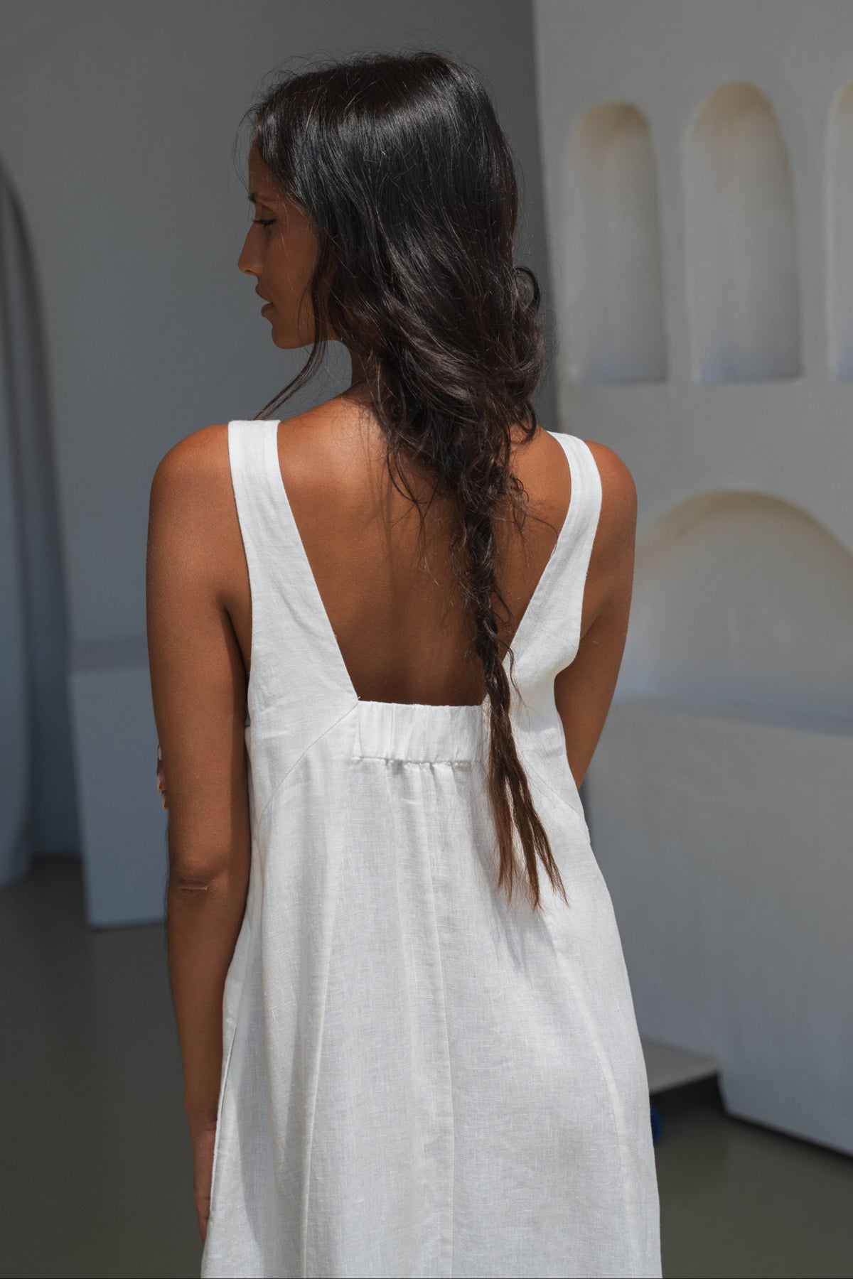 Woman in an ivory white LILLY PILLY Iris dress showing the back design view in a minimalistic room with arches.