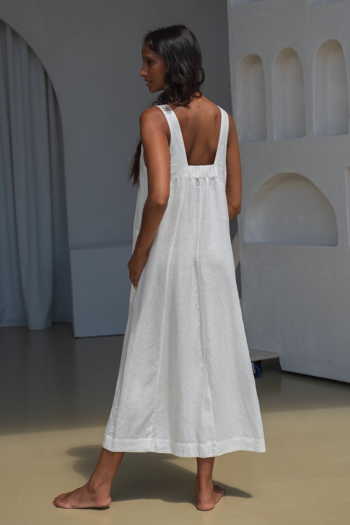LILLY PILLY woman showing the back view of an ivory white Iris dress standing  in a minimalistic room with white walls and a gray floor.