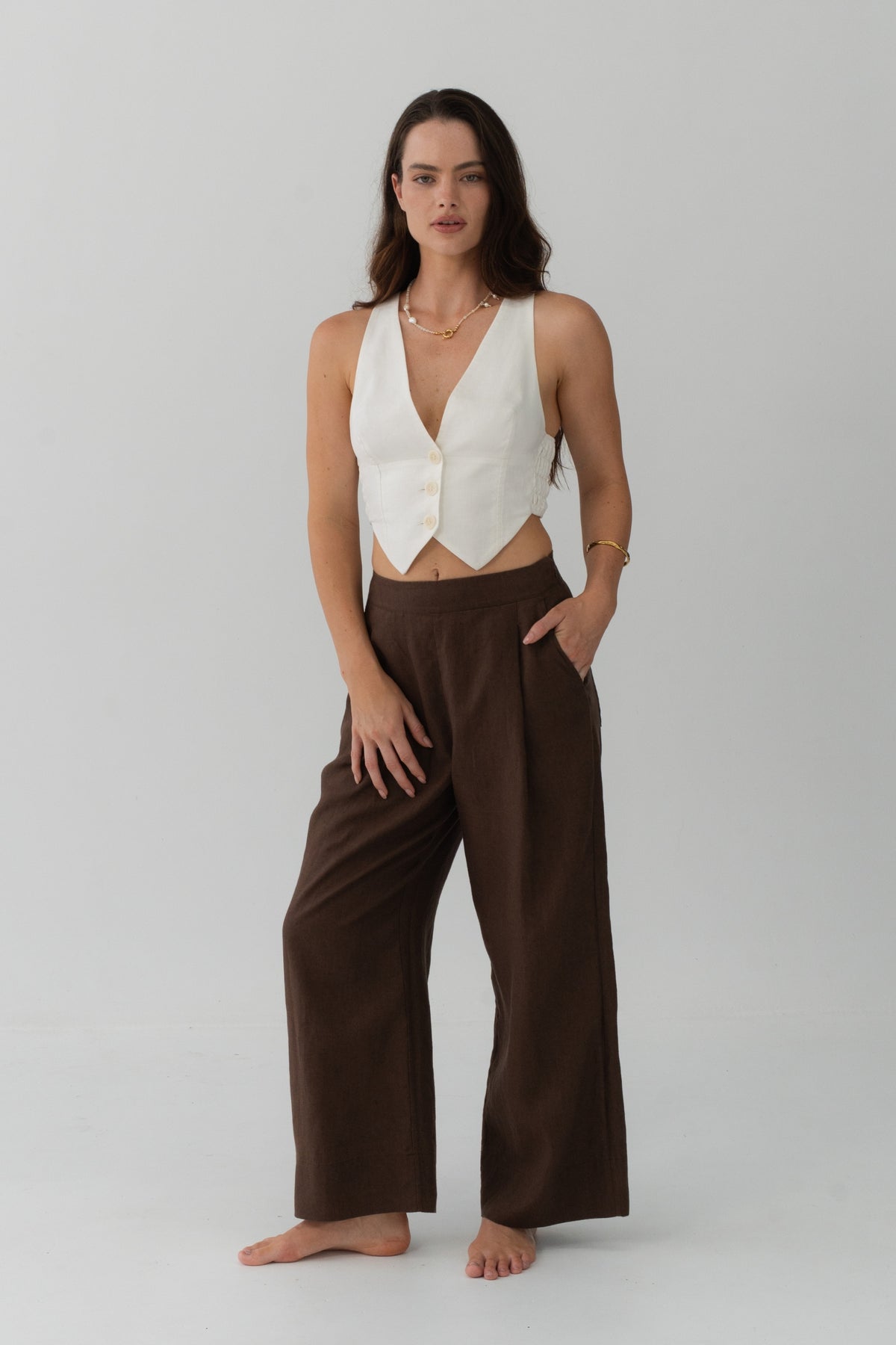 Ivy Linen Crop Pants in Chocolate - full length front view, woman wearing Chocolate wide-leg linen pants with ivory linen waistcoat, hand in pocket, studio backdrop - Lilly Pilly Collection