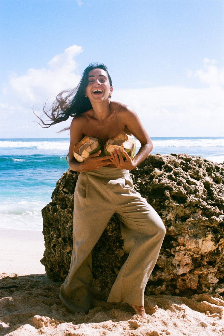 Ivy Linen Crop Pants in Taupe - woman sitting on coastal rocks wearing Taupe wide-leg linen crop pants, holding a coconuts, ocean backdrop - Lilly Pilly Collection