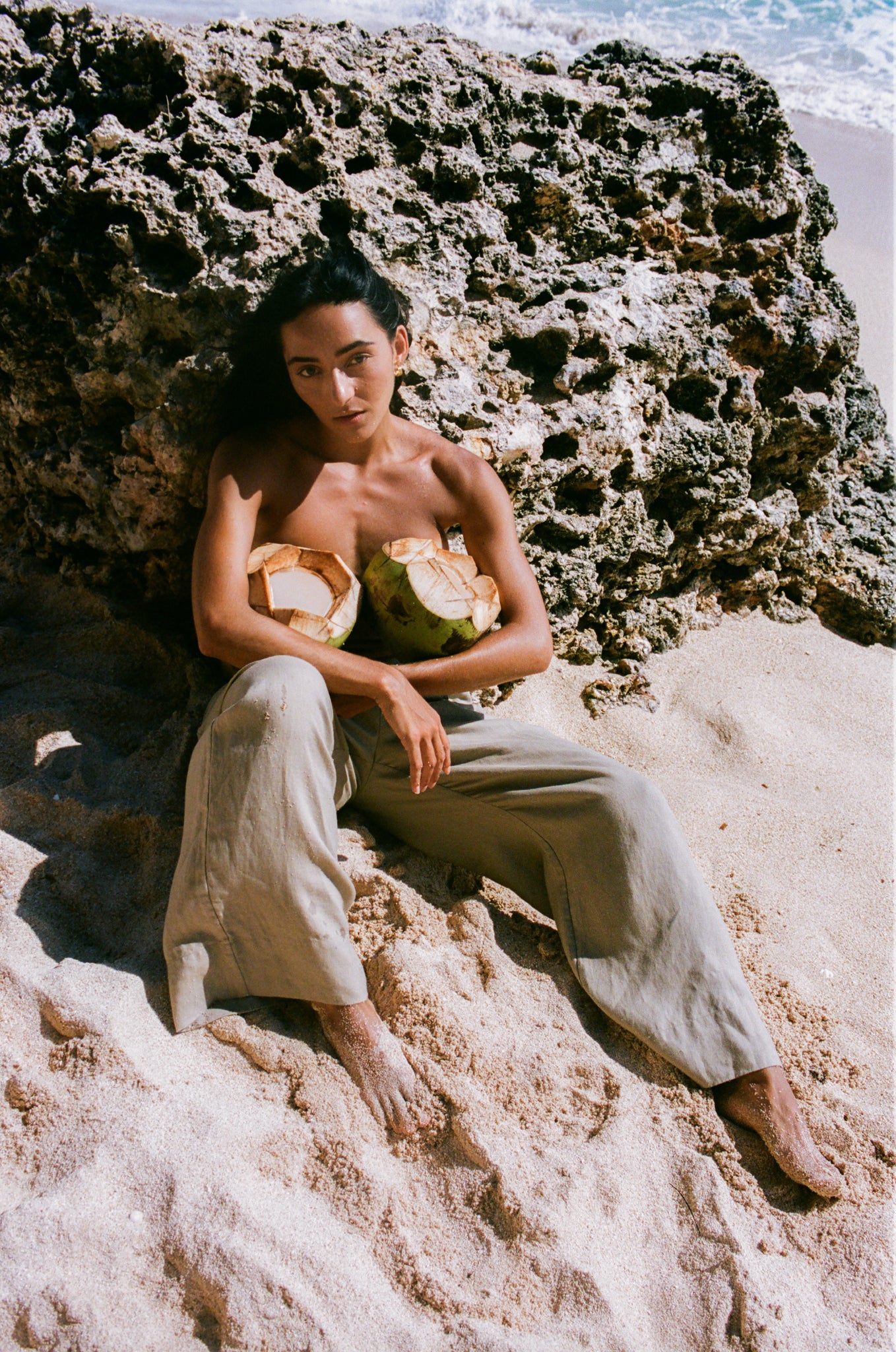 LILLY PILLY woman wearing  Ivy linen pants sitting on a rocky beach holding coconuts